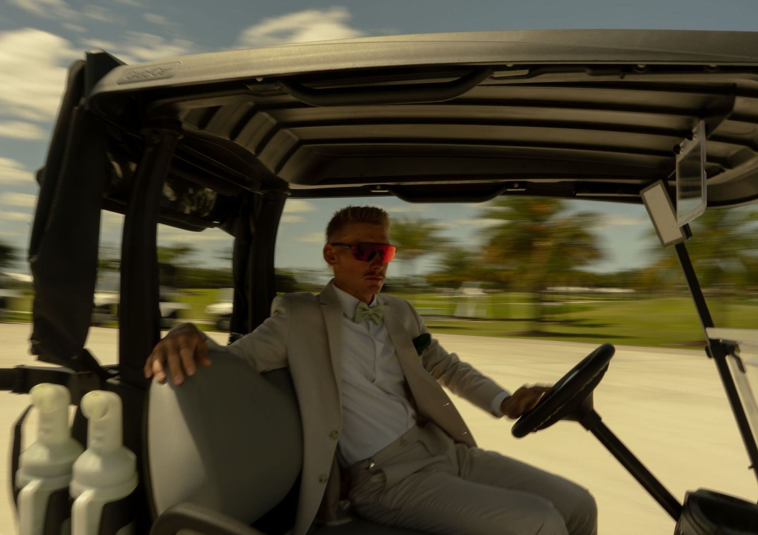 Stylish man in suit and sunglasses driving a golf cart on a sunny day at a tropical golf course.