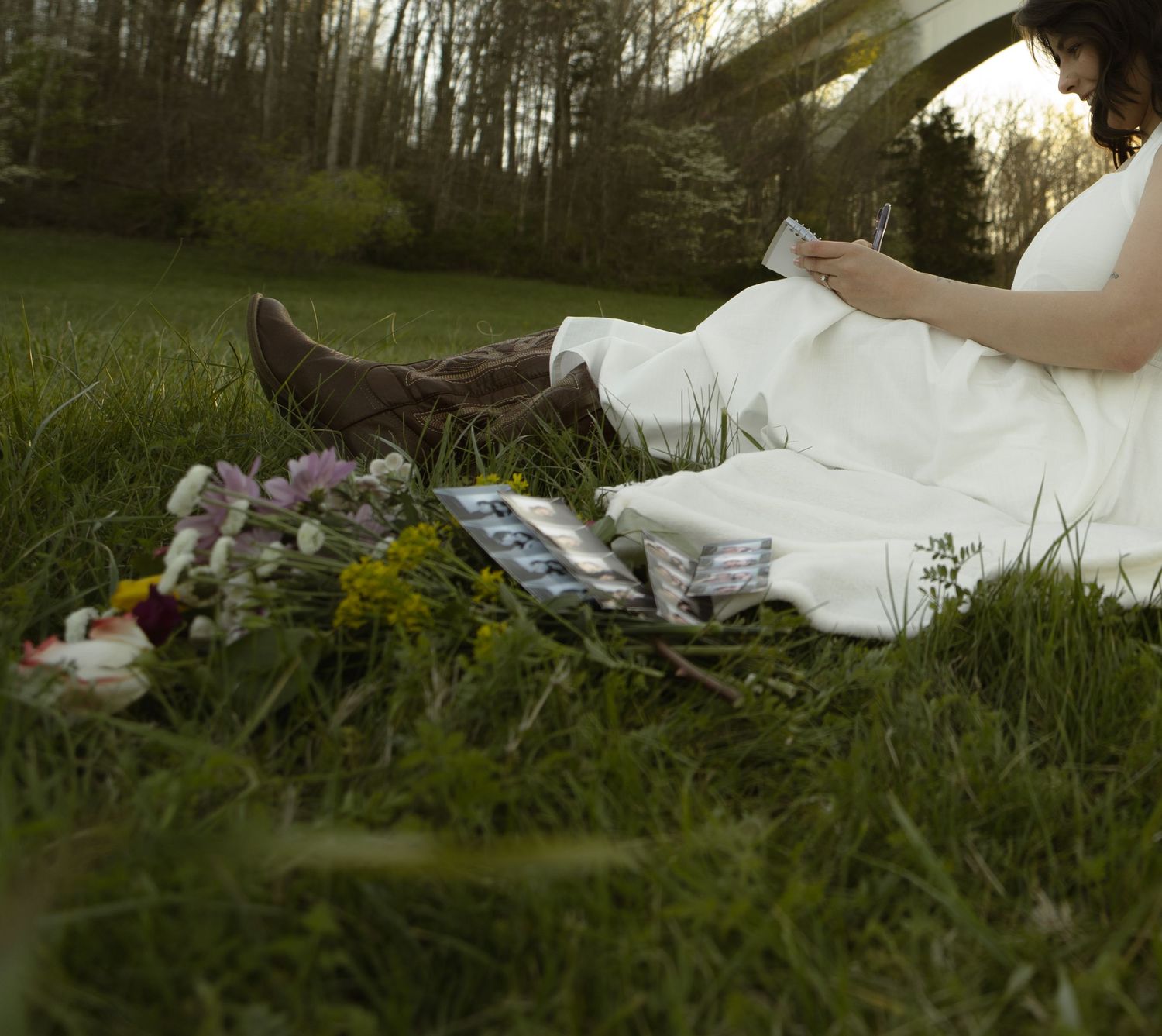 Pregnant woman in white dress and brown boots sitting on grass reading, with flowers and photos beside her.