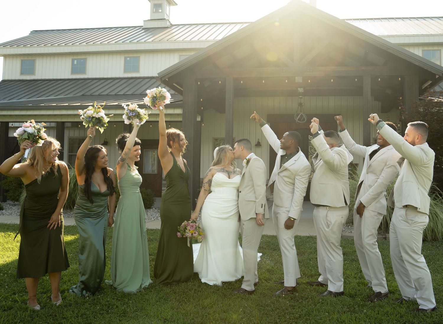 Wedding party celebrating outdoors, bride and groom kissing while bridesmaids in green dresses and groomsmen in tan suits cheer.