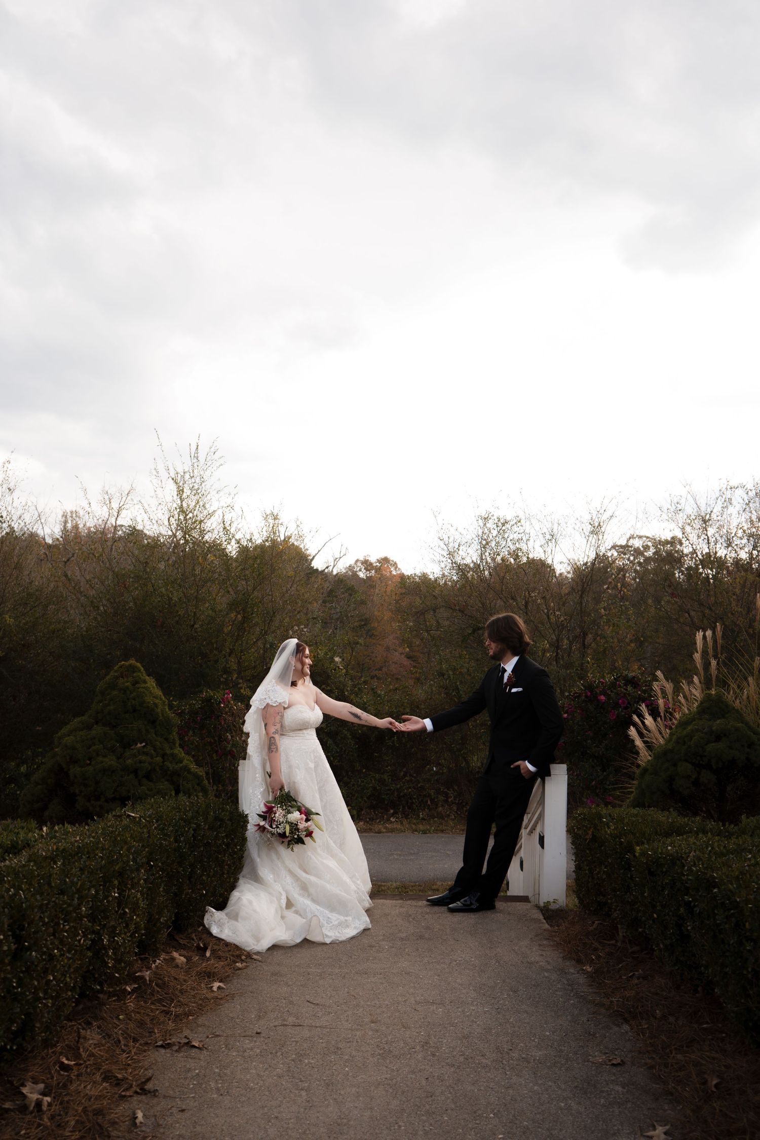 A couple in wedding attire stands facing each other on a garden path surrounded by autumn trees on an overcast day.