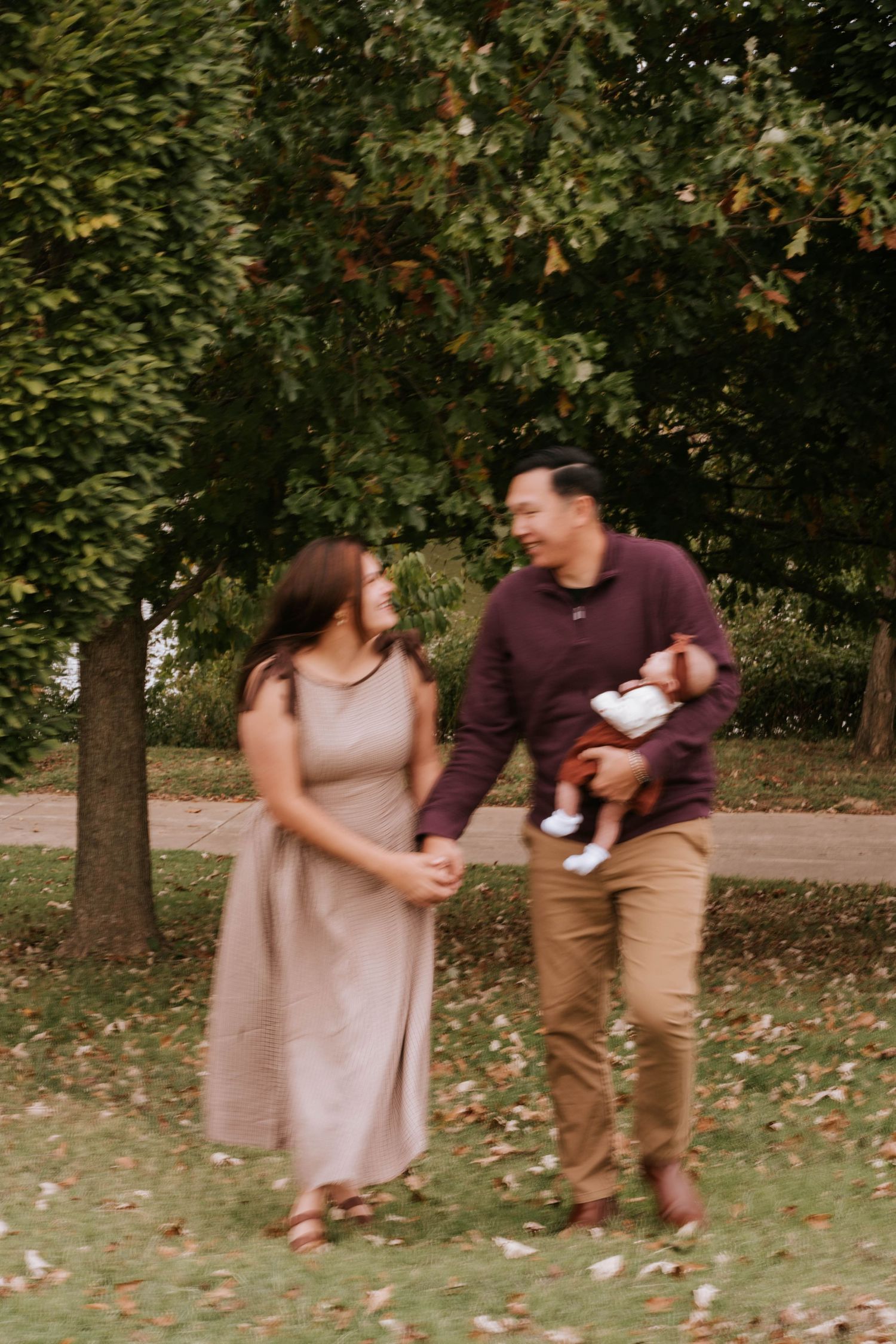 A couple in casual attire playfully interacts in a park setting with autumn leaves on the ground.