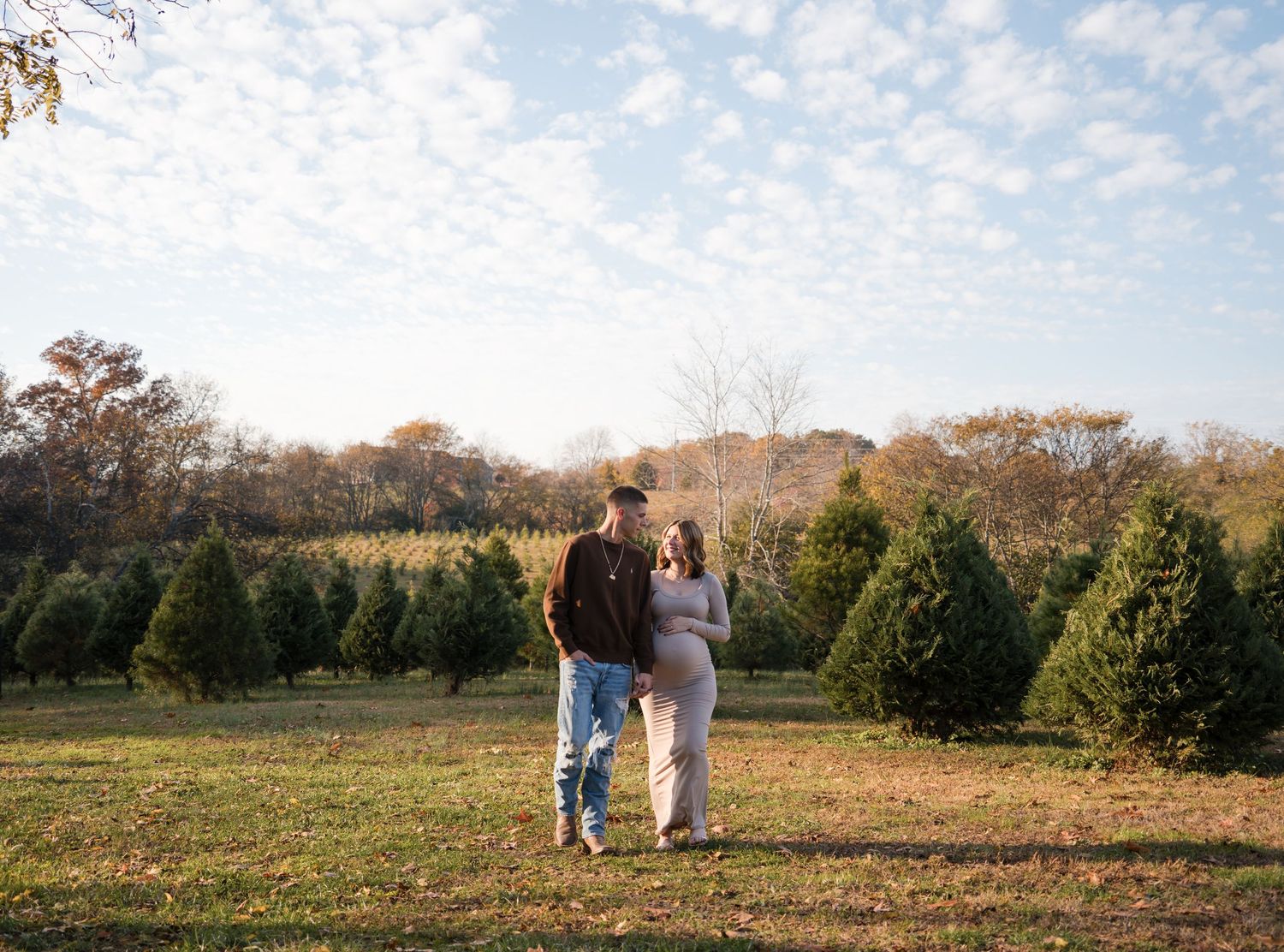 A couple walks through a Christmas tree farm on a sunny autumn afternoon with rows of evergreens in the background.
