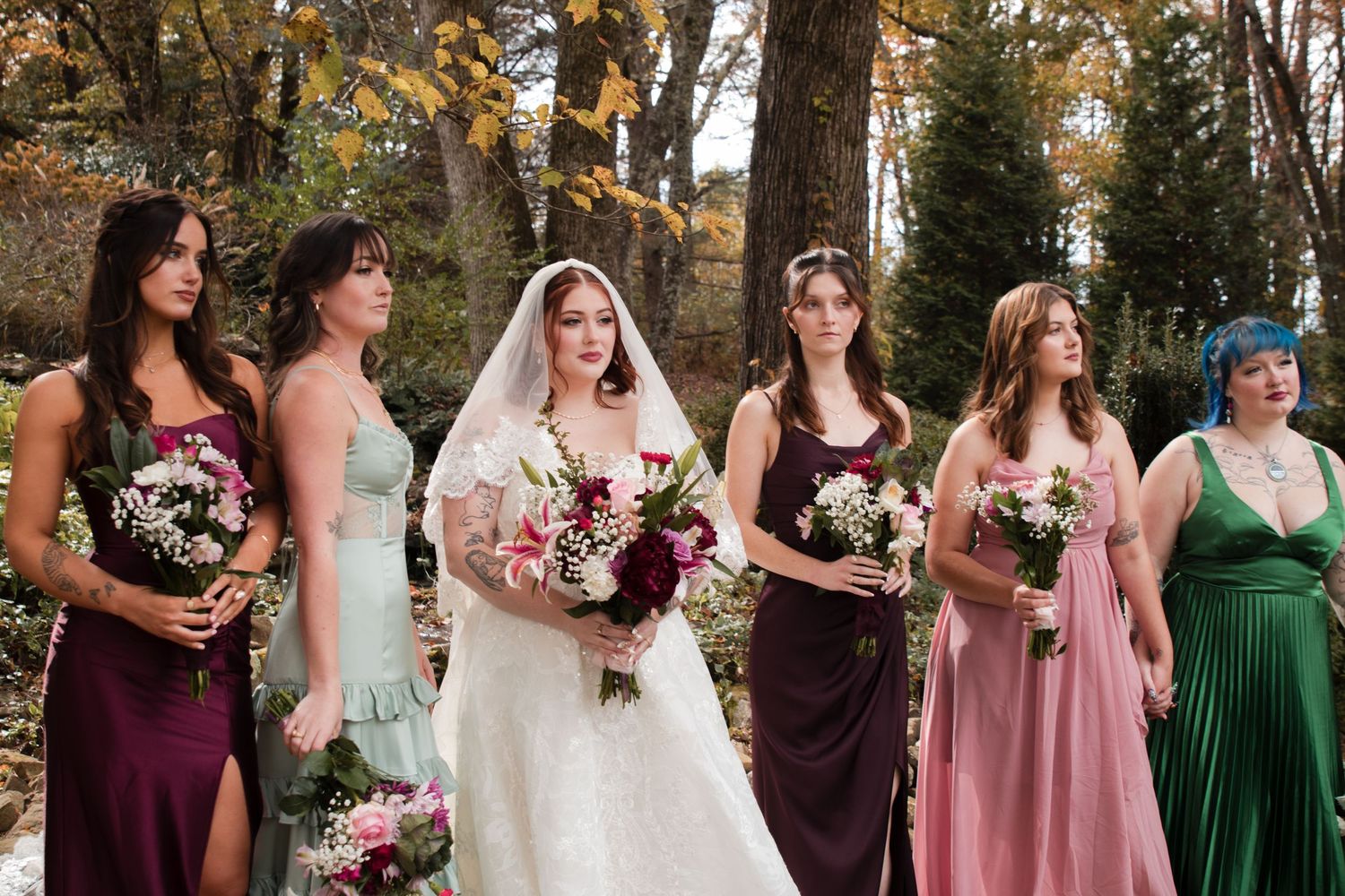 Wedding party stands together in autumn setting wearing burgundy, pink, and emerald dresses with floral bouquets.