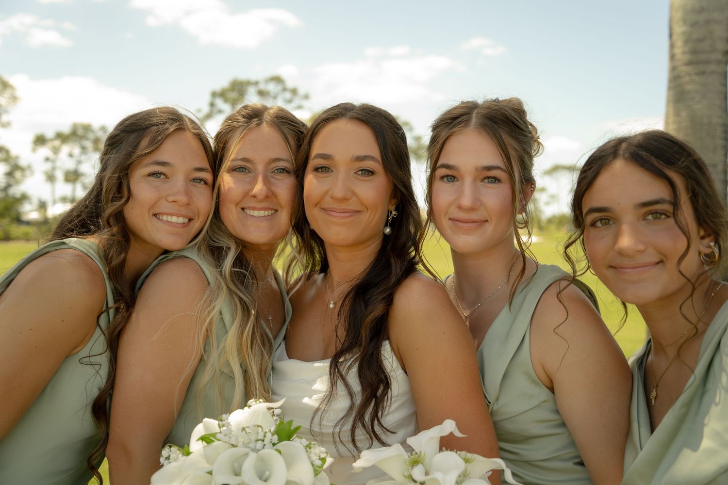 Smiling bride holding white bouquet poses with four bridesmaids in sage green dresses outdoors.