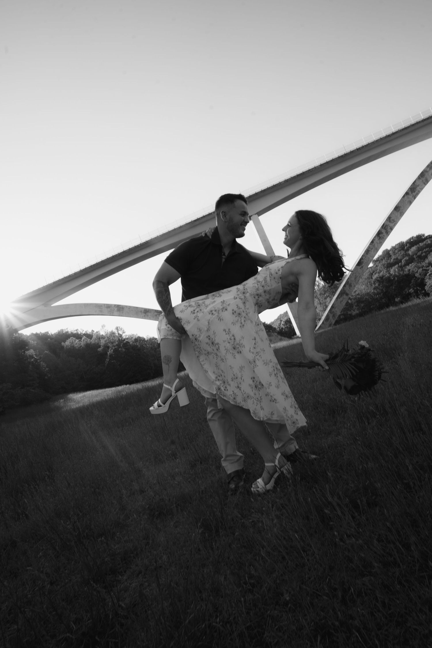 Black and white photo of a couple dancing and laughing together in a field with an arch bridge at sunset.