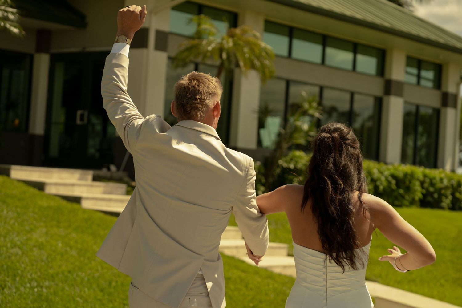 A bride and groom celebrate joyfully outside a elegant venue, with the groom raising his fist in triumph.