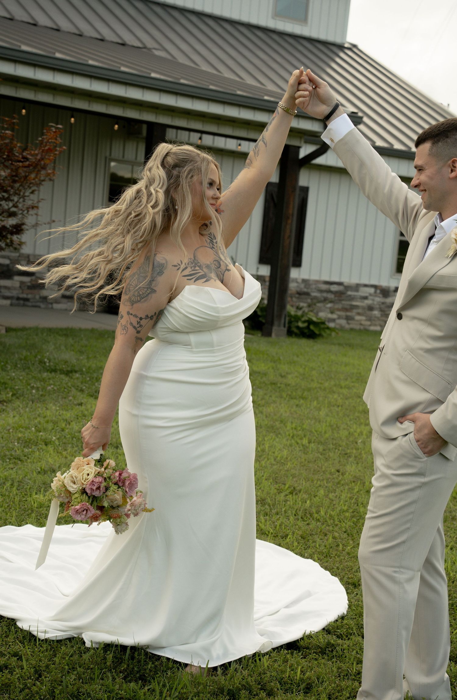 Bride in white gown twirling with groom in tan suit outdoors at barn venue, holding floral bouquet.