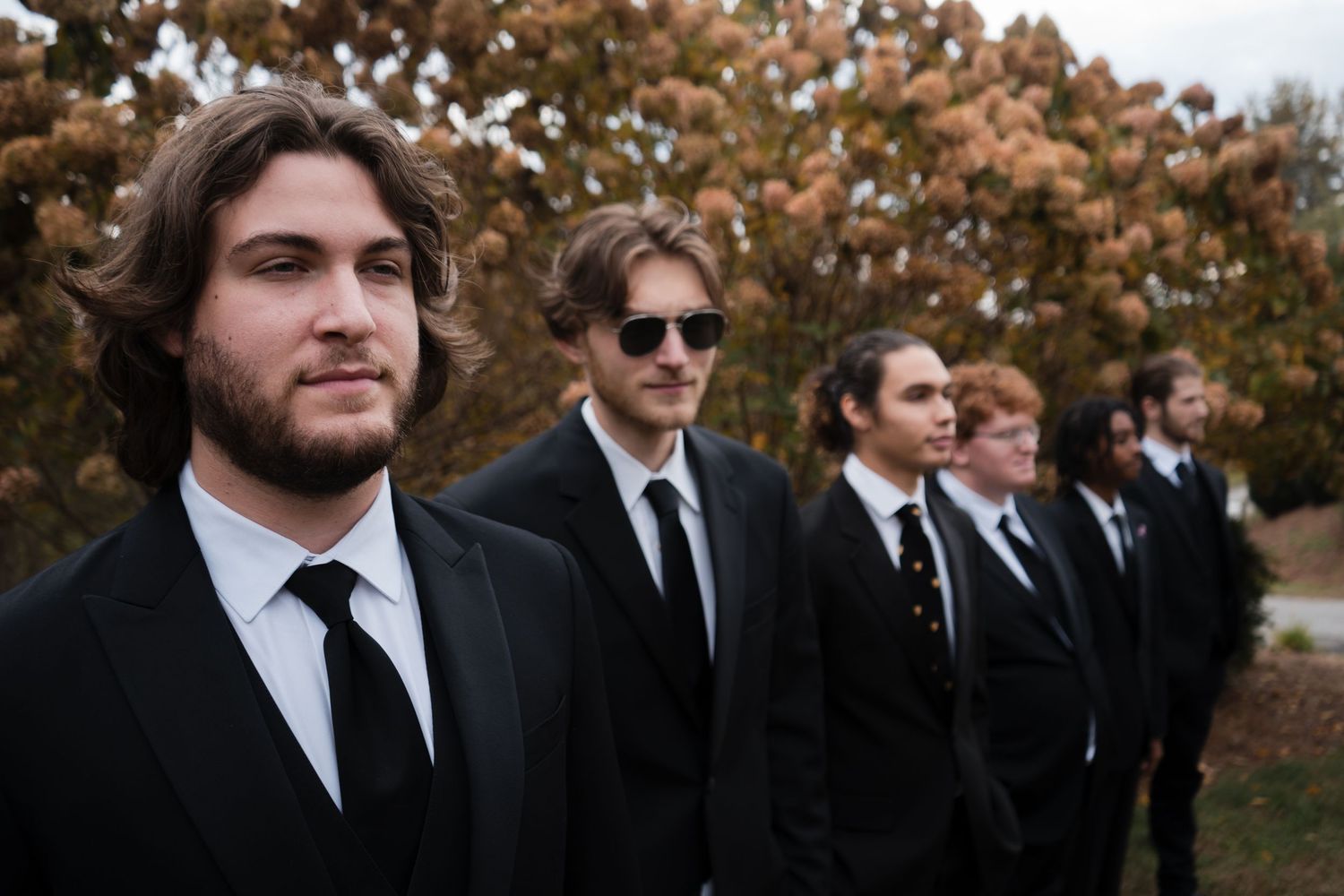 Groomsmen in black suits and ties stand in a line outdoors with autumn foliage in the background.