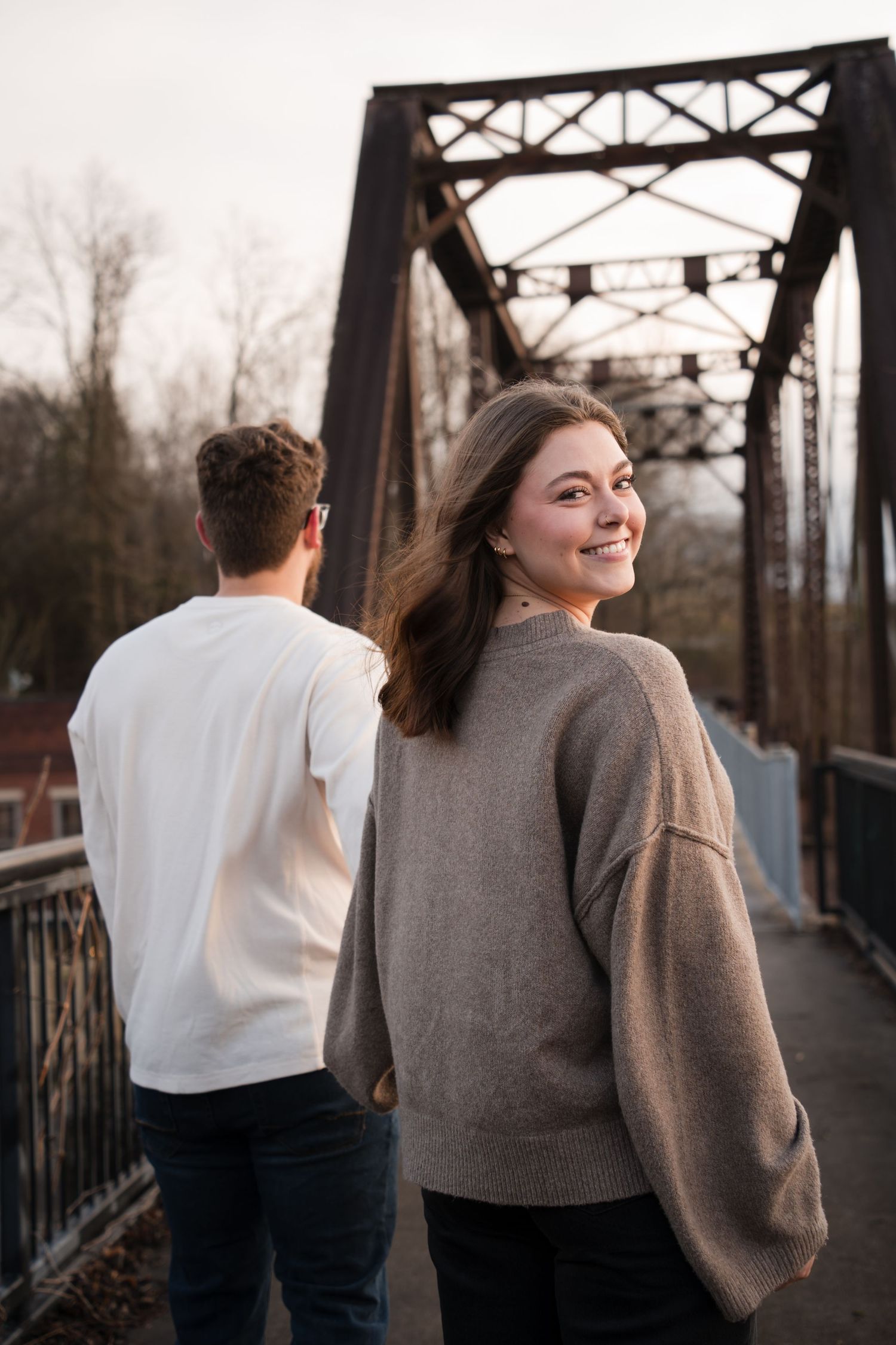 Young woman in gray sweater smiles over shoulder on iron bridge, man in white shirt beside her.