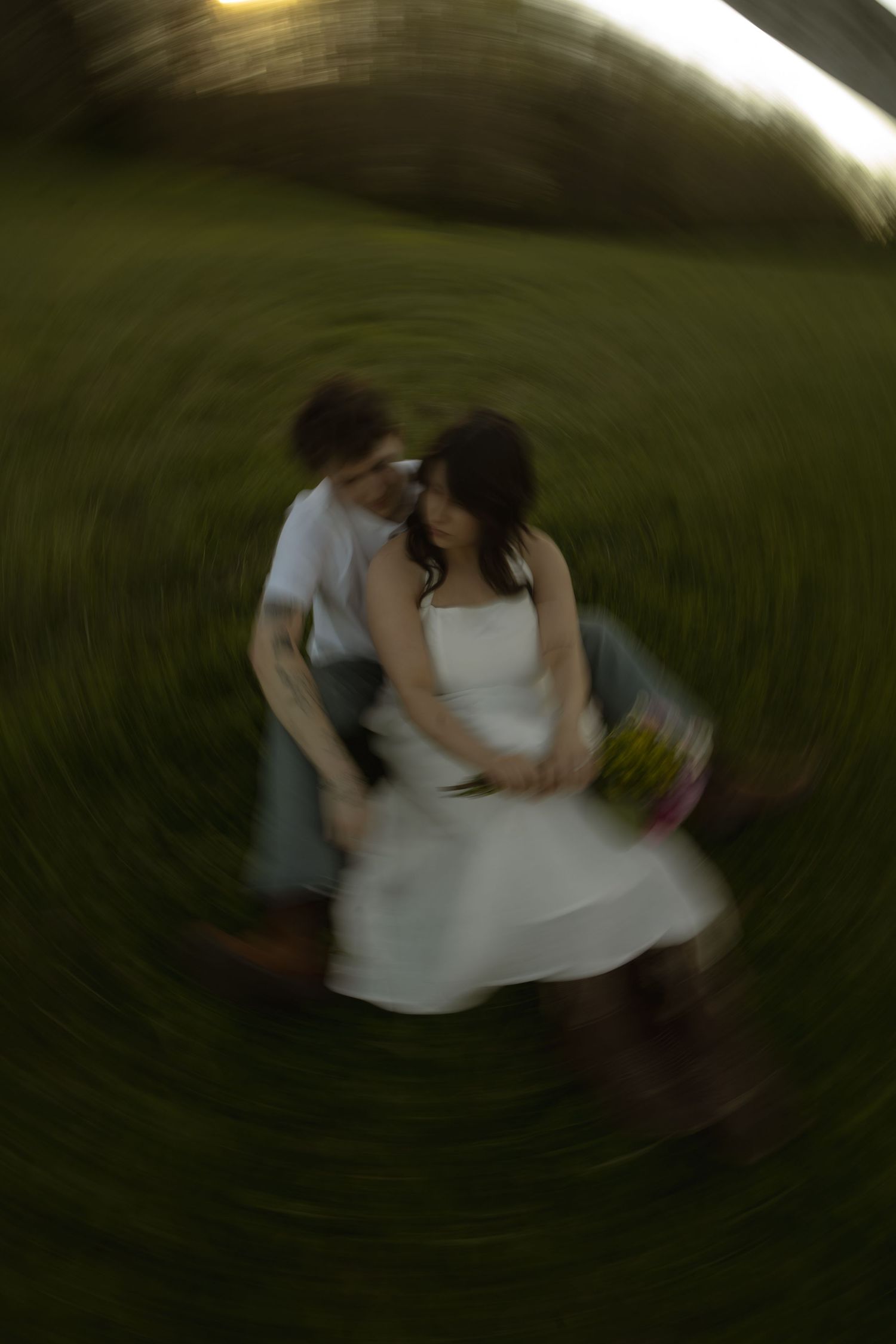 Couple embracing in a field, captured with a dreamy spin blur effect, bride in white dress holding flowers.