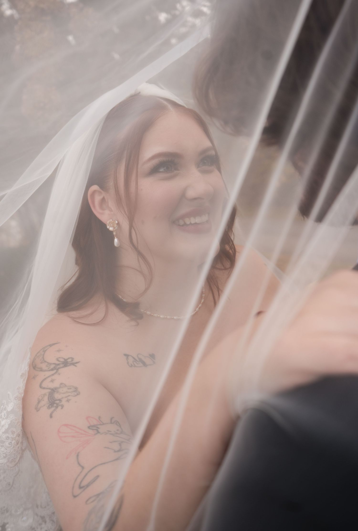 A bride wearing a white veil smiles radiantly through soft focus with visible tattoos on her shoulder.