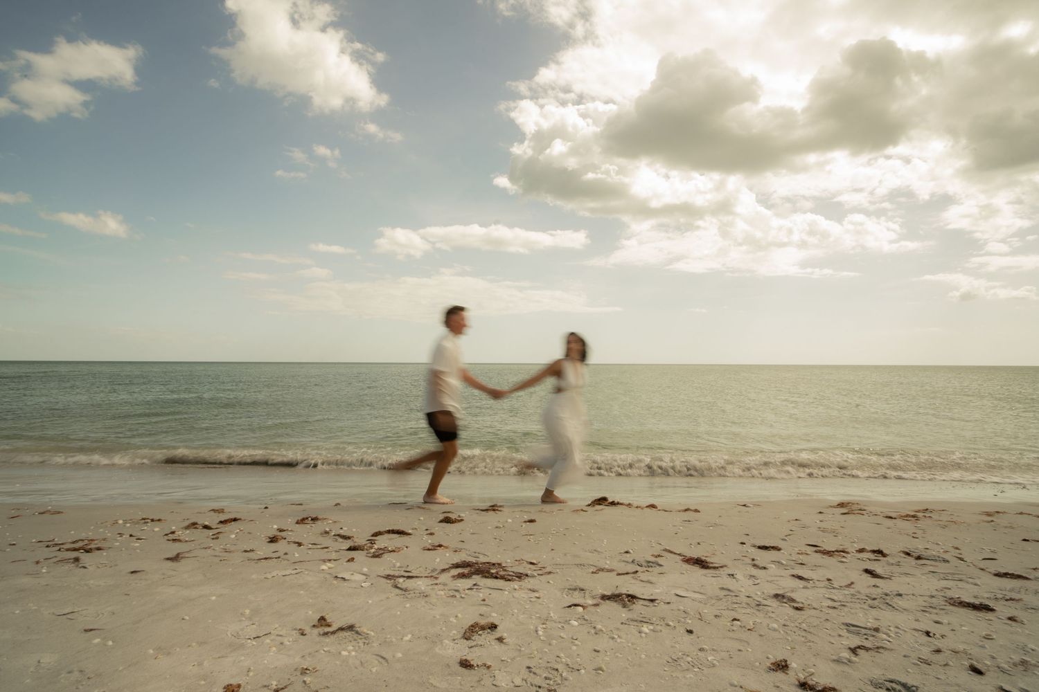A couple joyfully runs hand-in-hand along a sandy beach with ocean waves and cloudy sky backdrop.