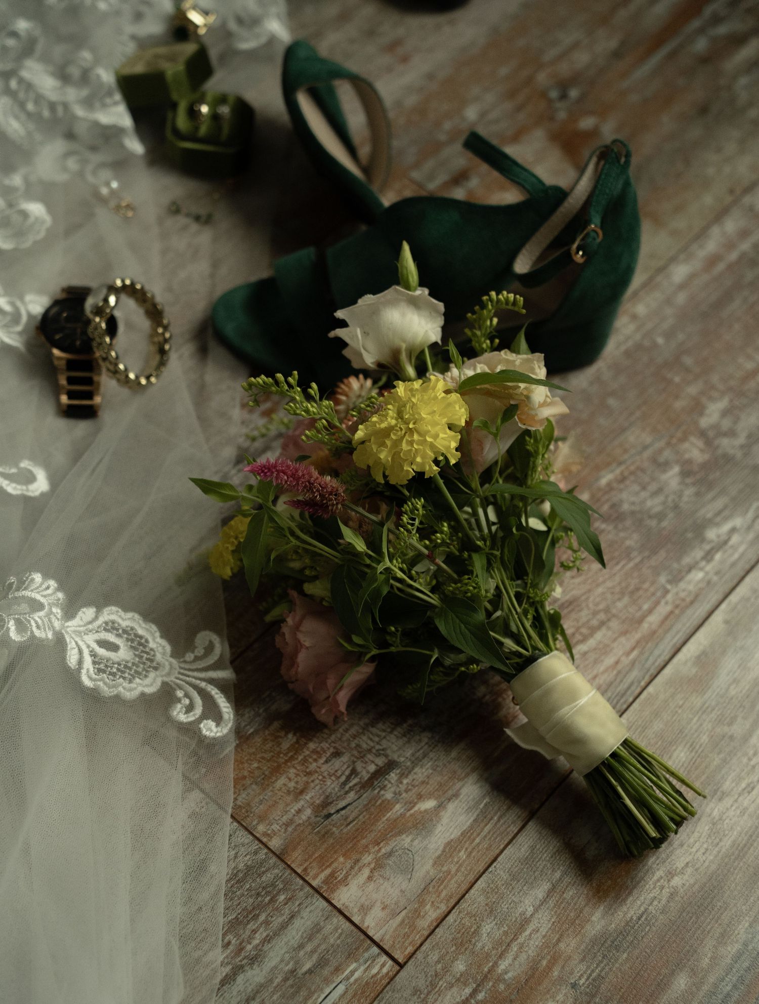 Bridal flat lay with wildflower bouquet, emerald green heels, lace veil, gold jewelry, and perfume on rustic wood floor.