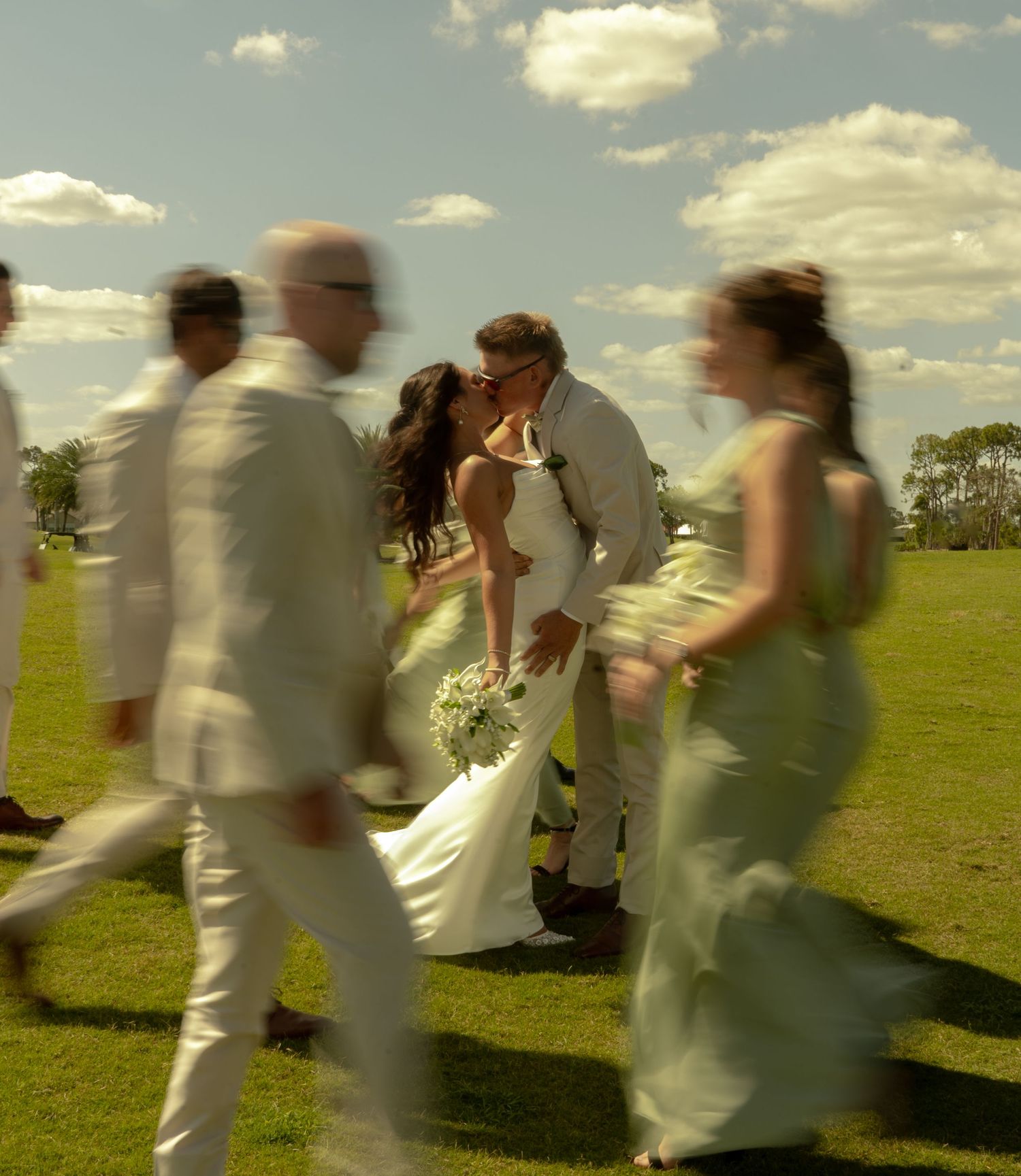 A bride and groom share a kiss surrounded by their wedding party on a sunny green lawn.
