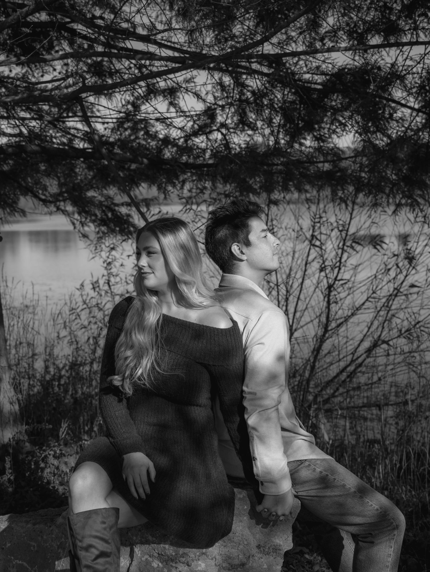 A romantic black and white portrait of a couple sitting back-to-back by a serene lake under overhanging tree branches.