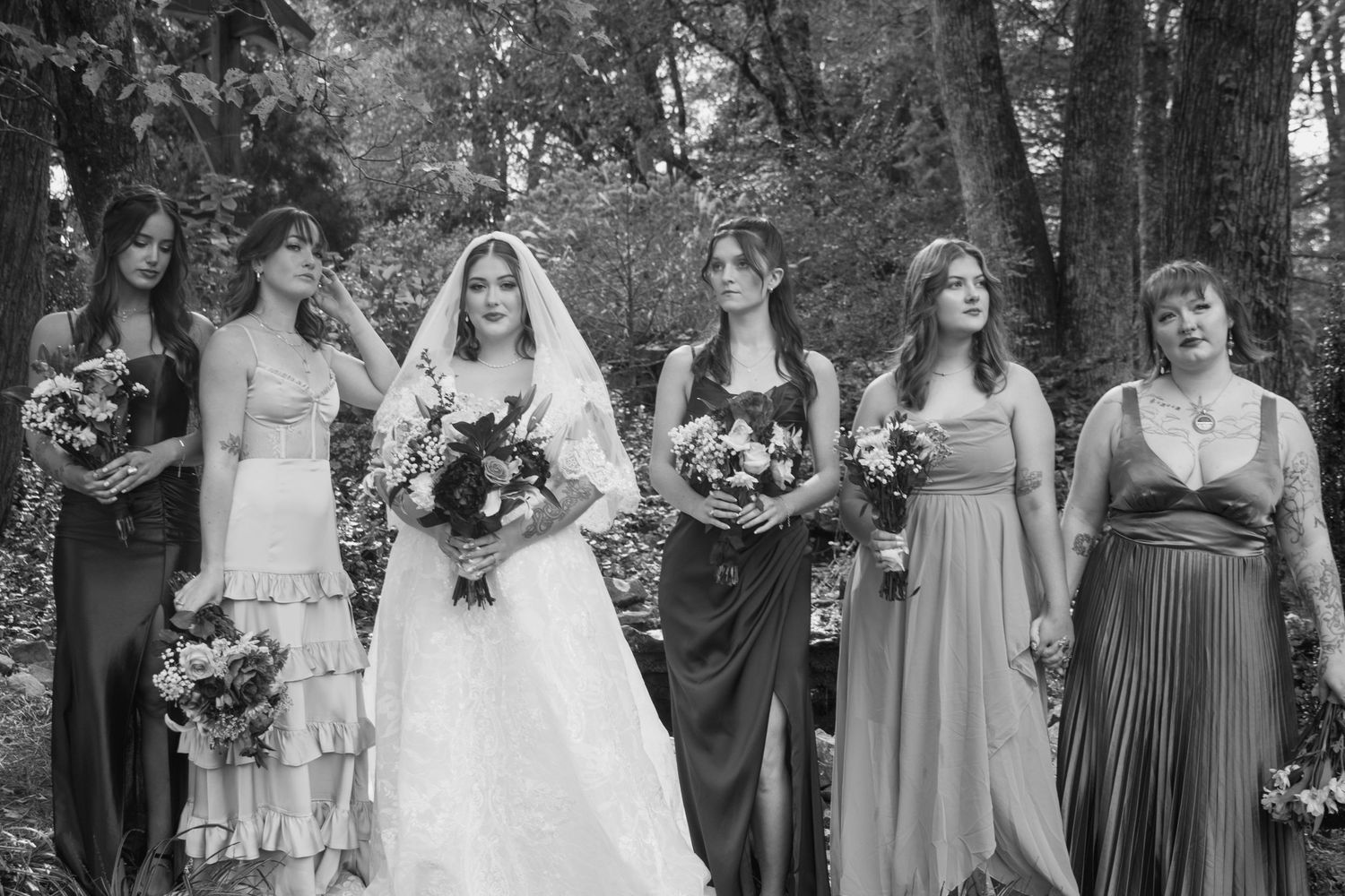Wedding party members in gray and white dresses pose together outdoors holding floral bouquets.