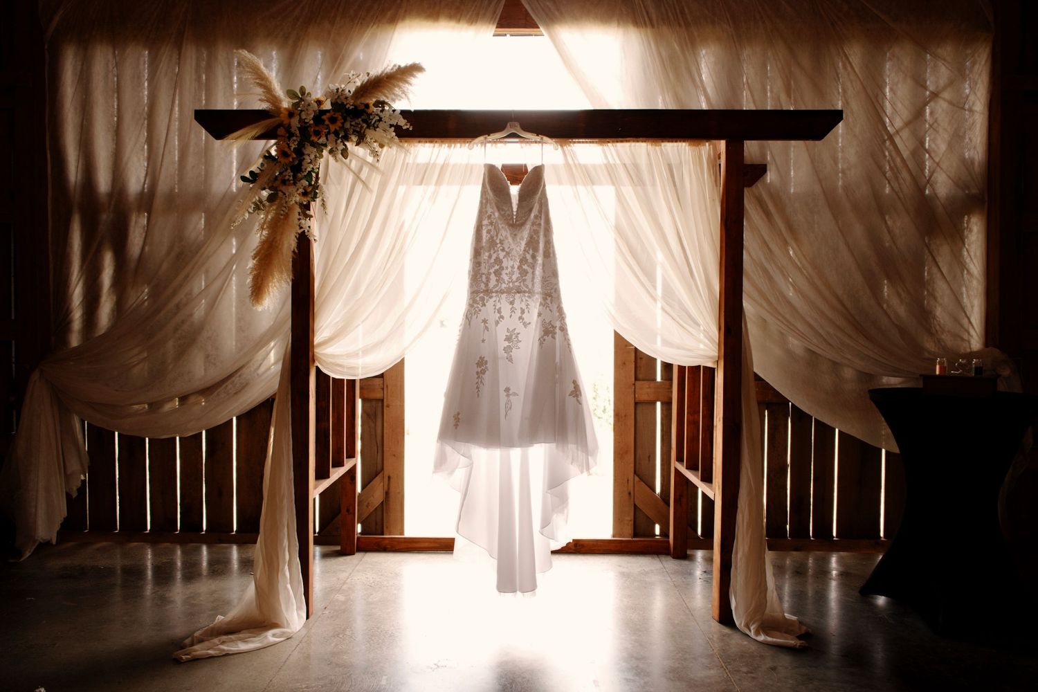 Ethereal white wedding dress hangs in rustic barn with soft natural light streaming through wooden slats.