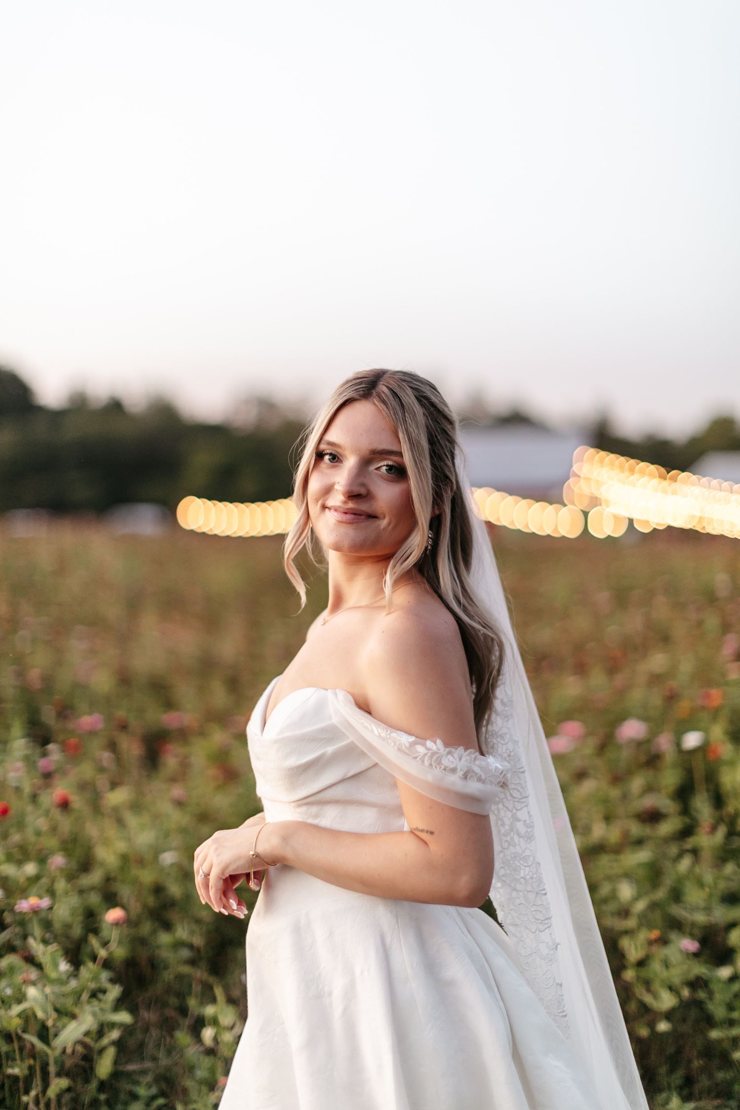 A bride in a flowing white dress poses in a golden-lit field during sunset.