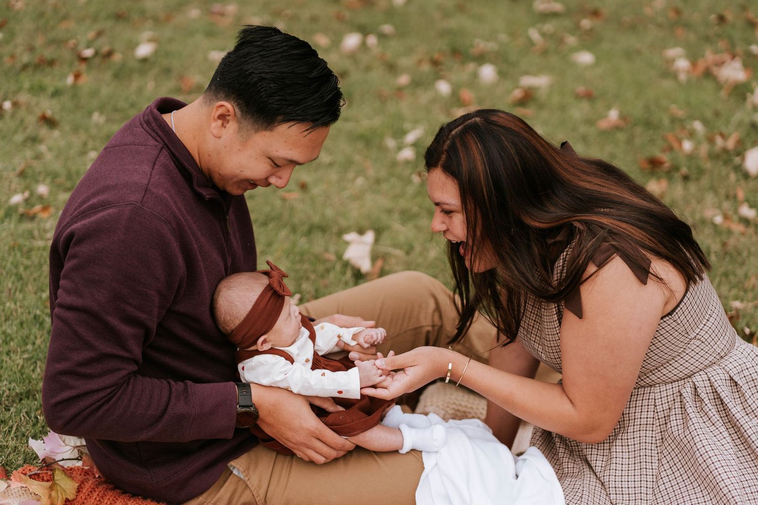 A couple sits on grass during autumn while tenderly interacting with a newborn baby on a blanket.