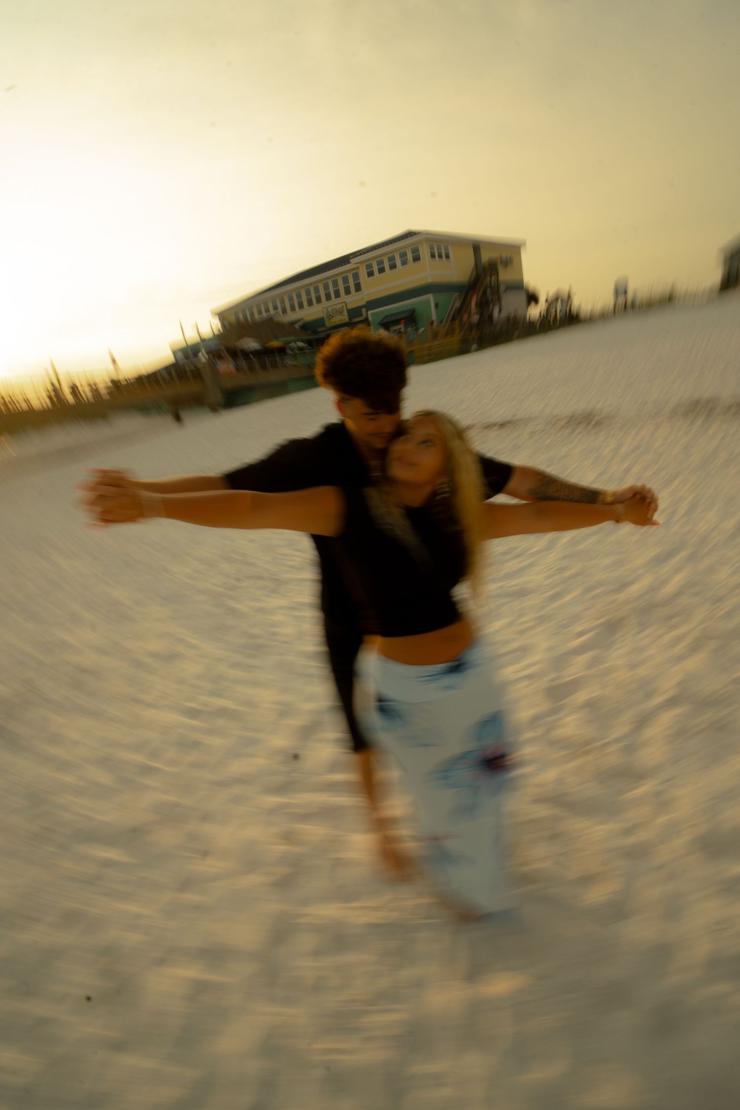 Couple dancing on a sandy beach at sunset with arms outstretched, blurred motion effect.
