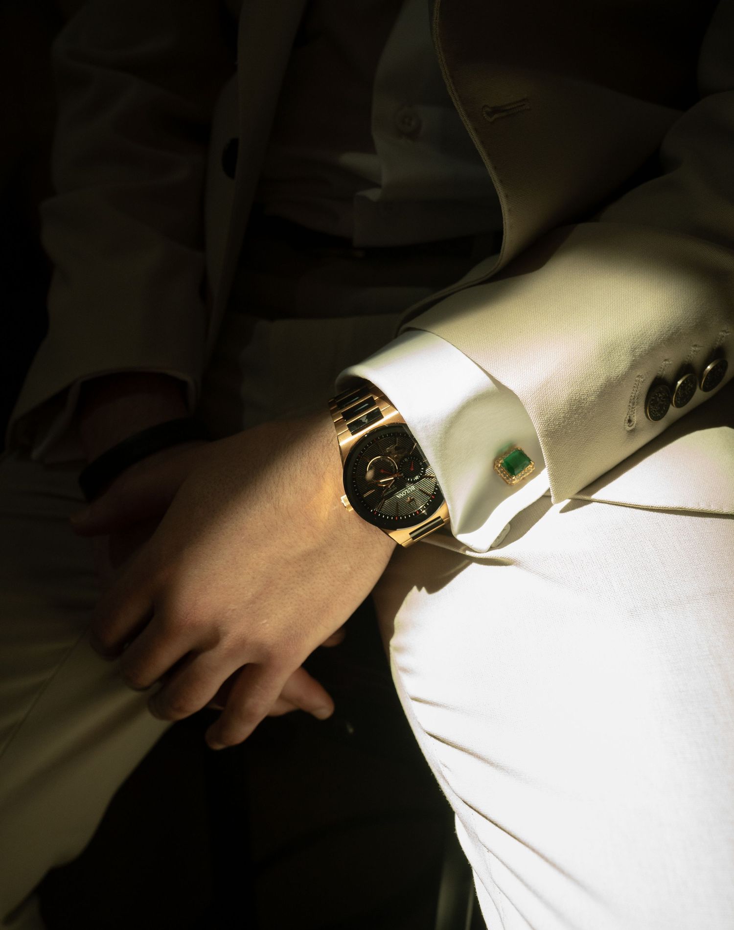 Person in white suit wearing a luxurious gold watch with green gem cufflink, elegant fashion accessory close-up.