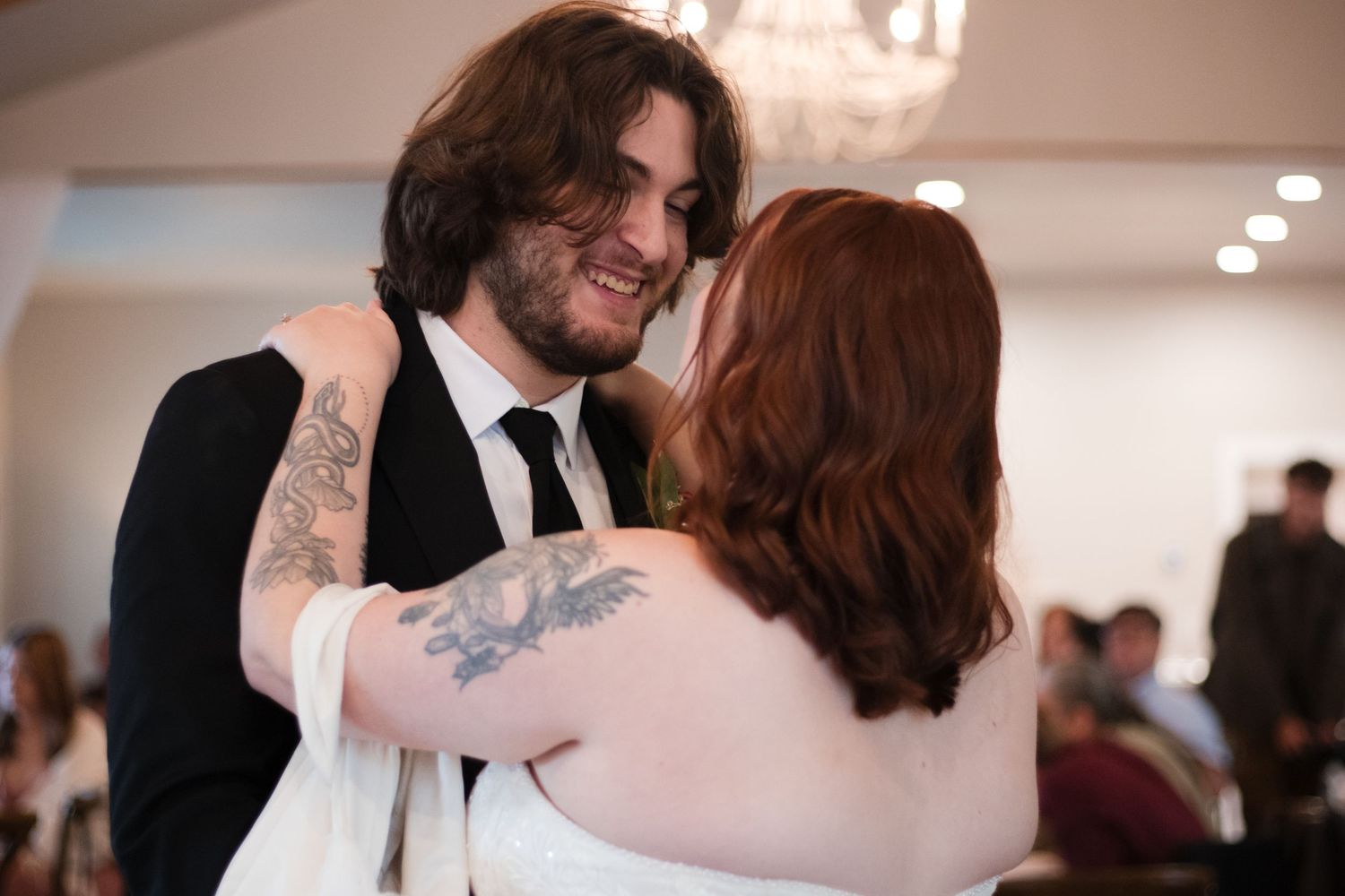 A couple shares a tender moment while dancing together at an indoor event, with the person in black wearing a suit and tie.