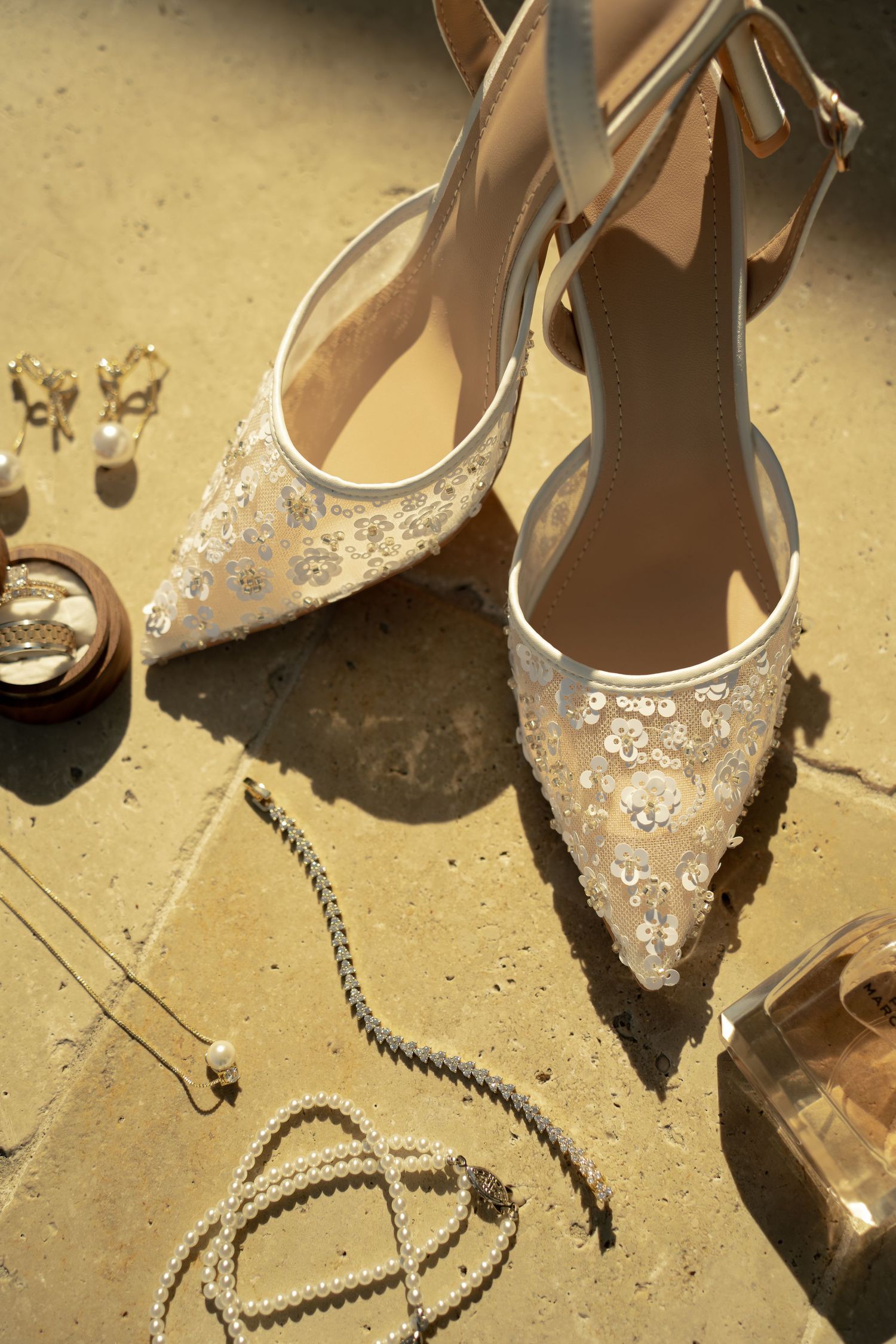 Elegant beaded bridal heels with ankle strap displayed alongside pearl jewelry and earrings on a stone surface.