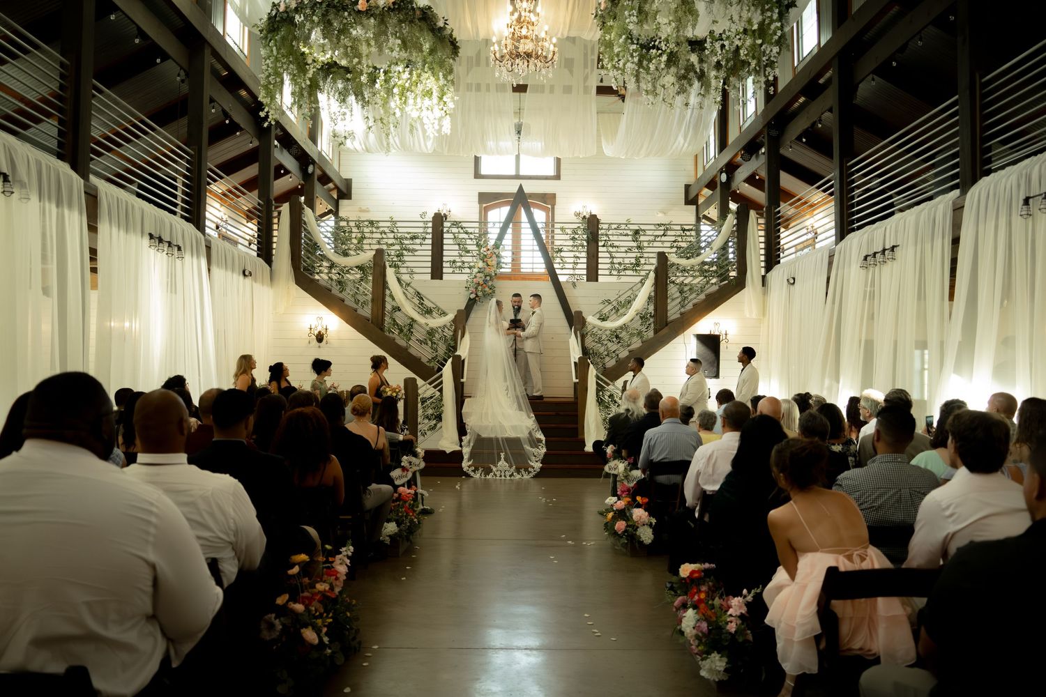 Elegant wedding ceremony in a rustic barn venue with floral arch, chandelier, and guests seated along the aisle.
