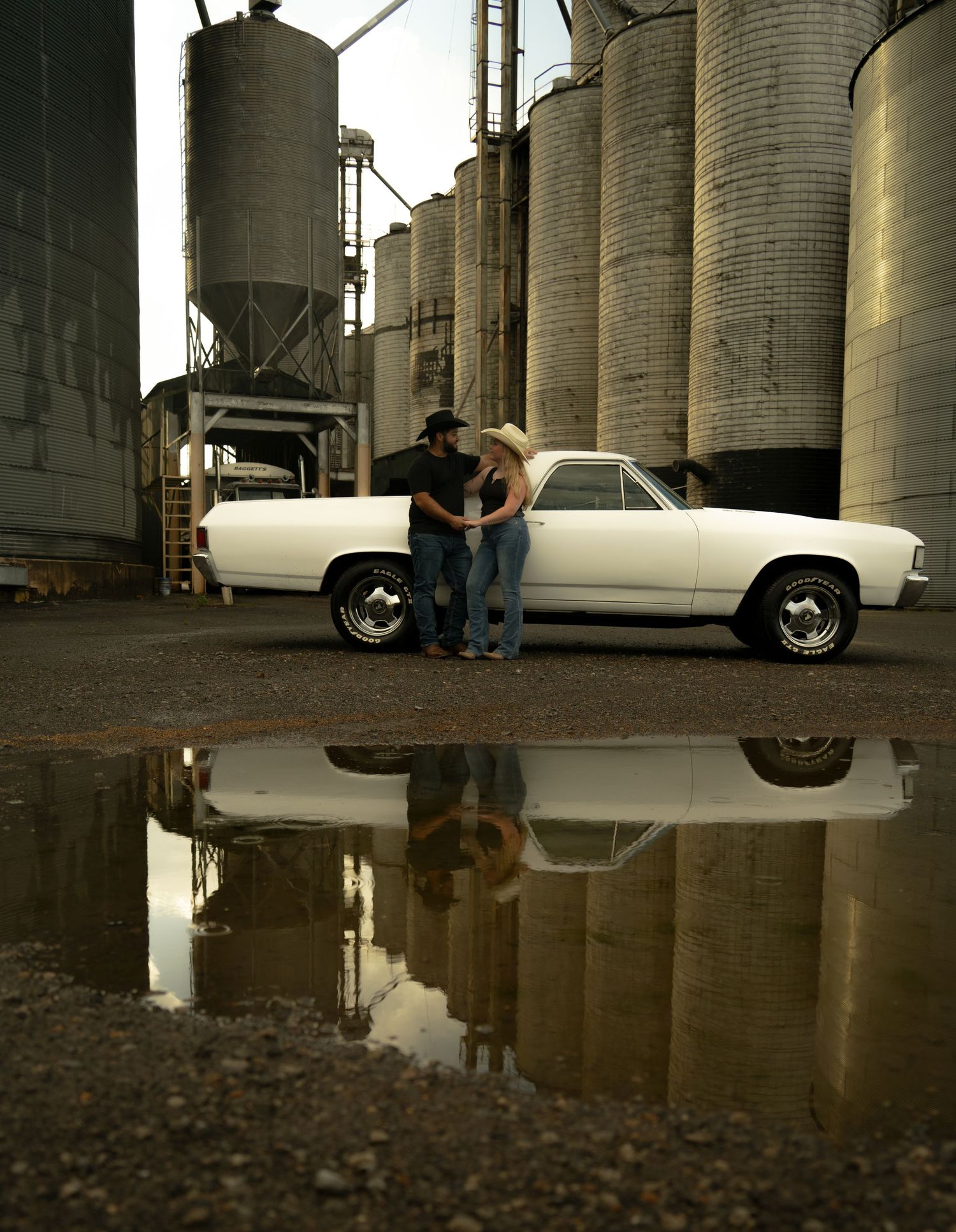 Couple in cowboy hats standing by a white classic car near grain silos, reflected in a puddle below.