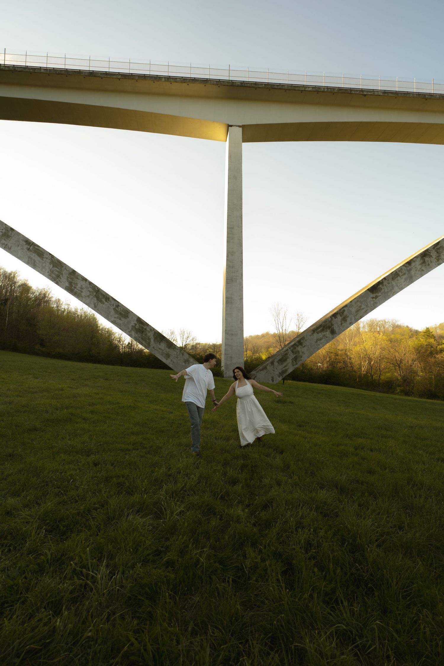 Couple in white holding hands under a large highway bridge on a green grassy field at sunset.