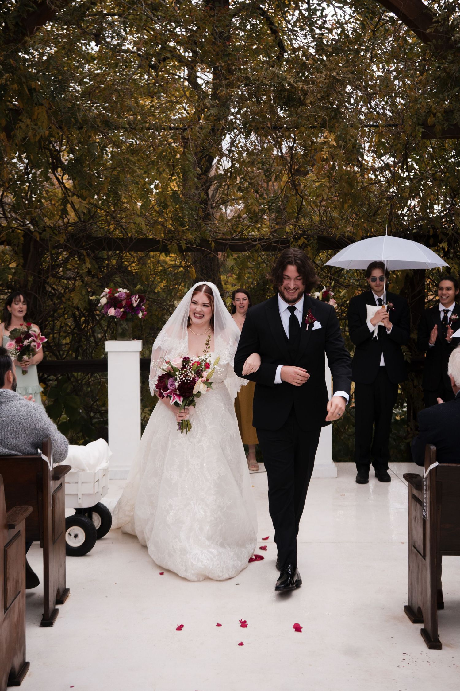 Wedding party members walk down an aisle decorated with rose petals in an outdoor ceremony setting.