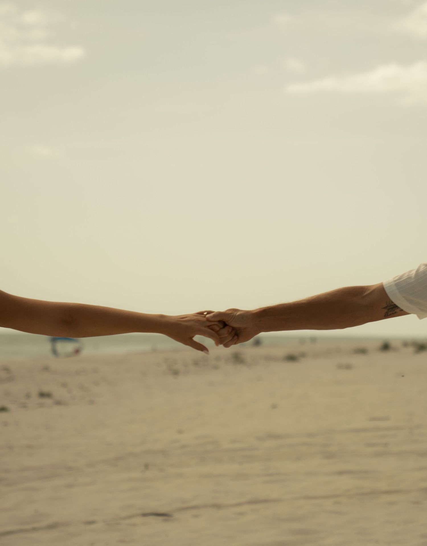 Two people holding hands on a sandy beach with a hazy sky backdrop.