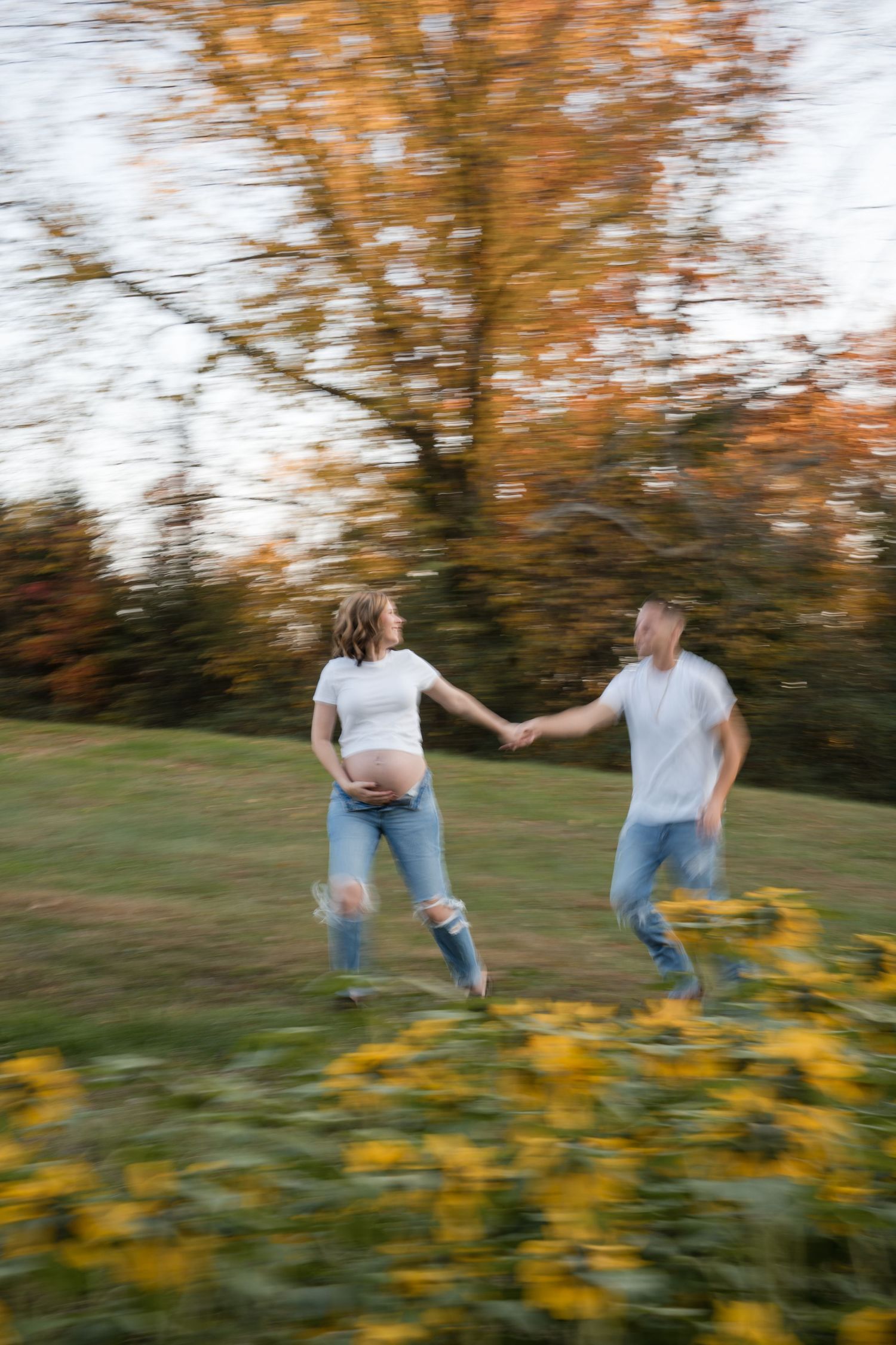 Two people run hand in hand through a field of yellow flowers with autumn trees in the background.