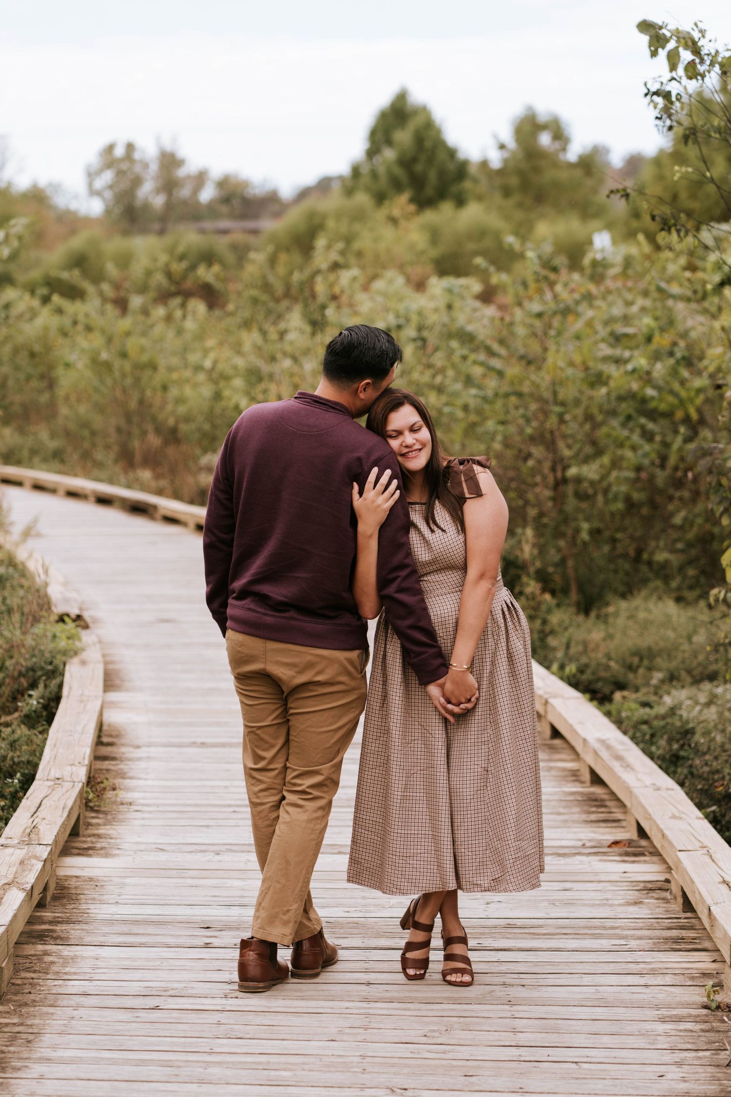 A couple walks and embraces on a wooden boardwalk surrounded by lush green vegetation on a sunny day.