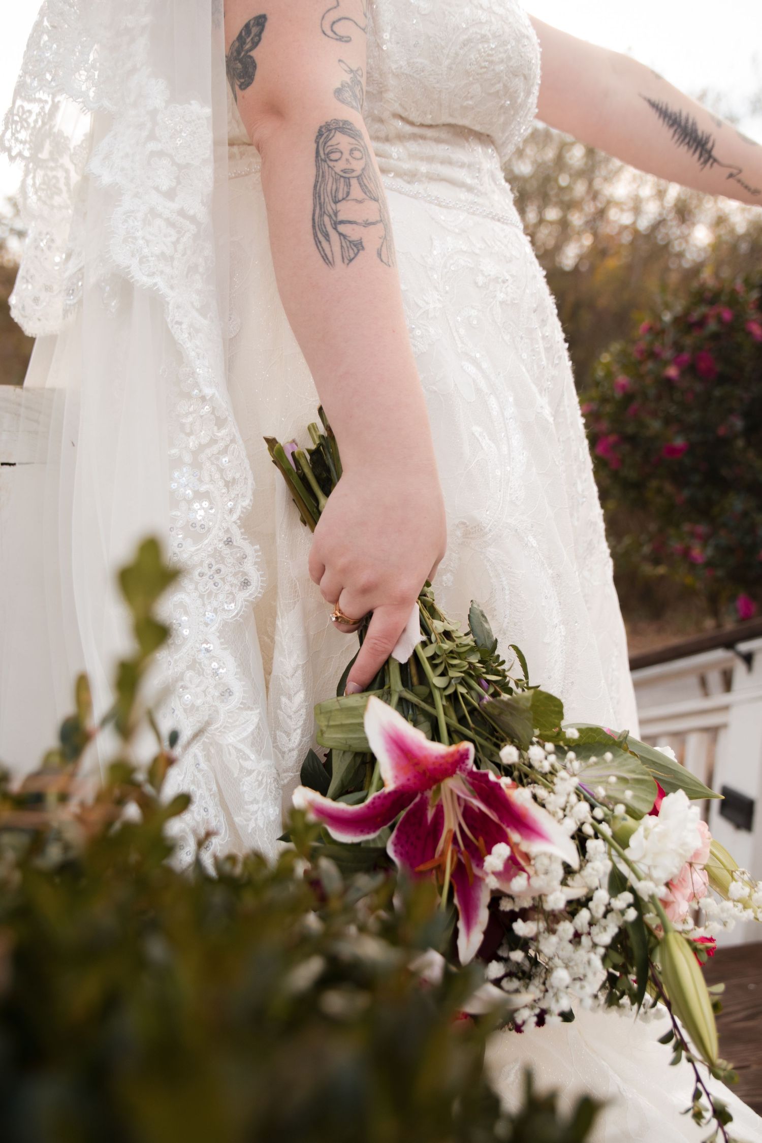 Close-up of a bride's tattooed arm holding a bouquet with pink lilies and lace dress details.