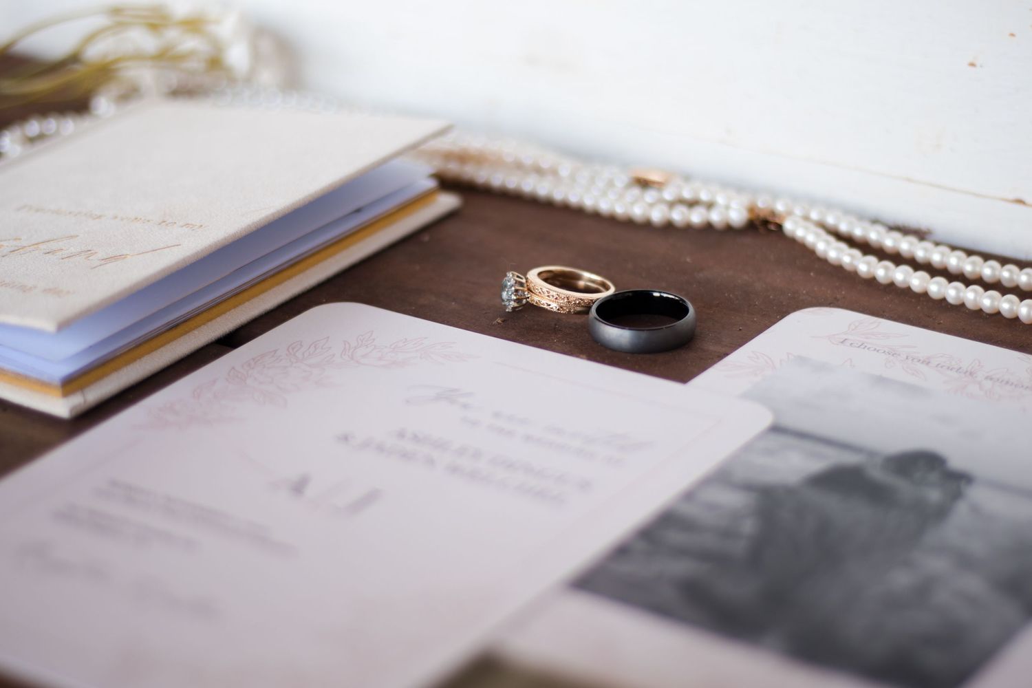 Wedding stationery and pearl necklace arranged on white surface with rings.