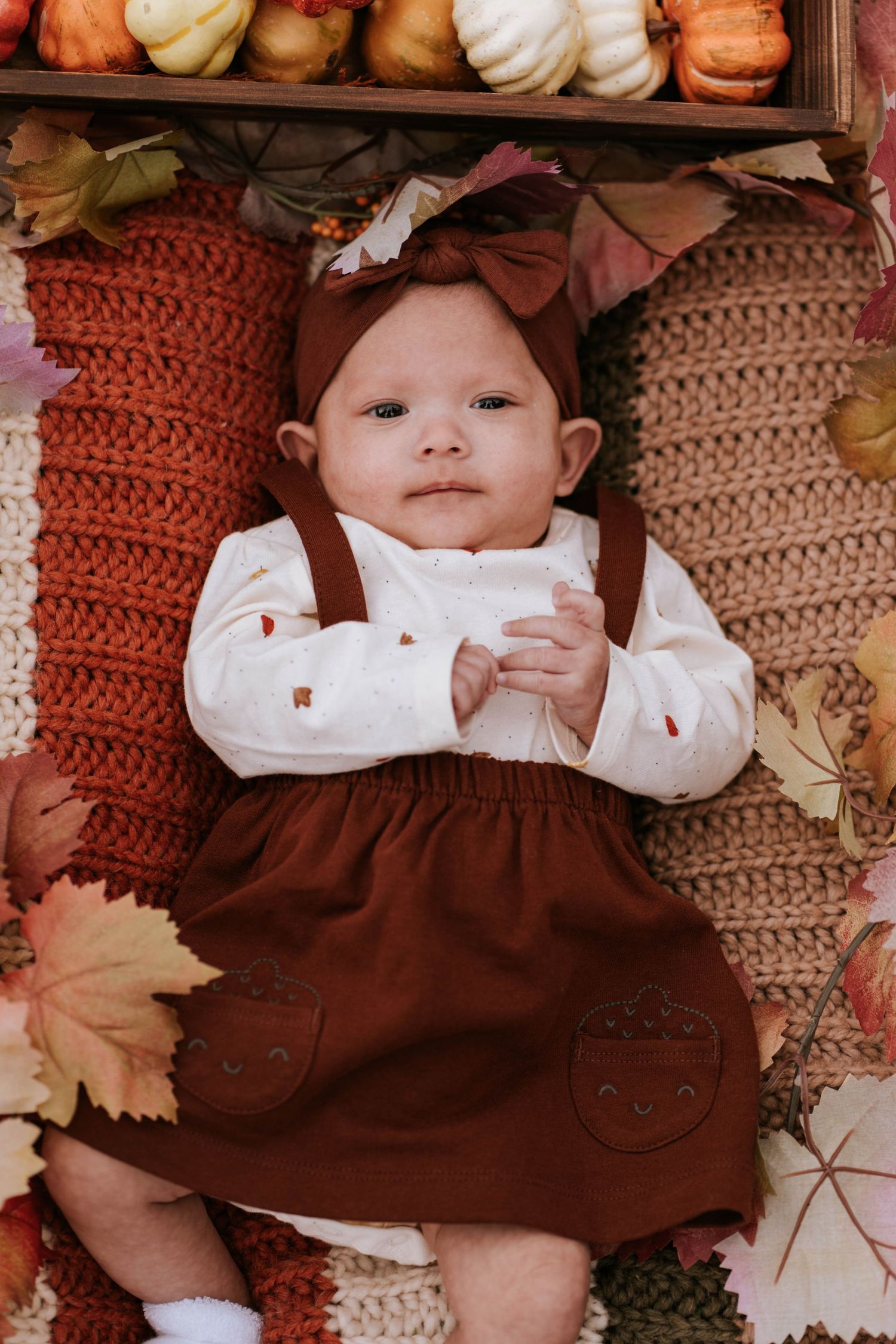 A small baby wearing a brown dress and white shirt lies among autumn decorations with pumpkins and knit pillows.