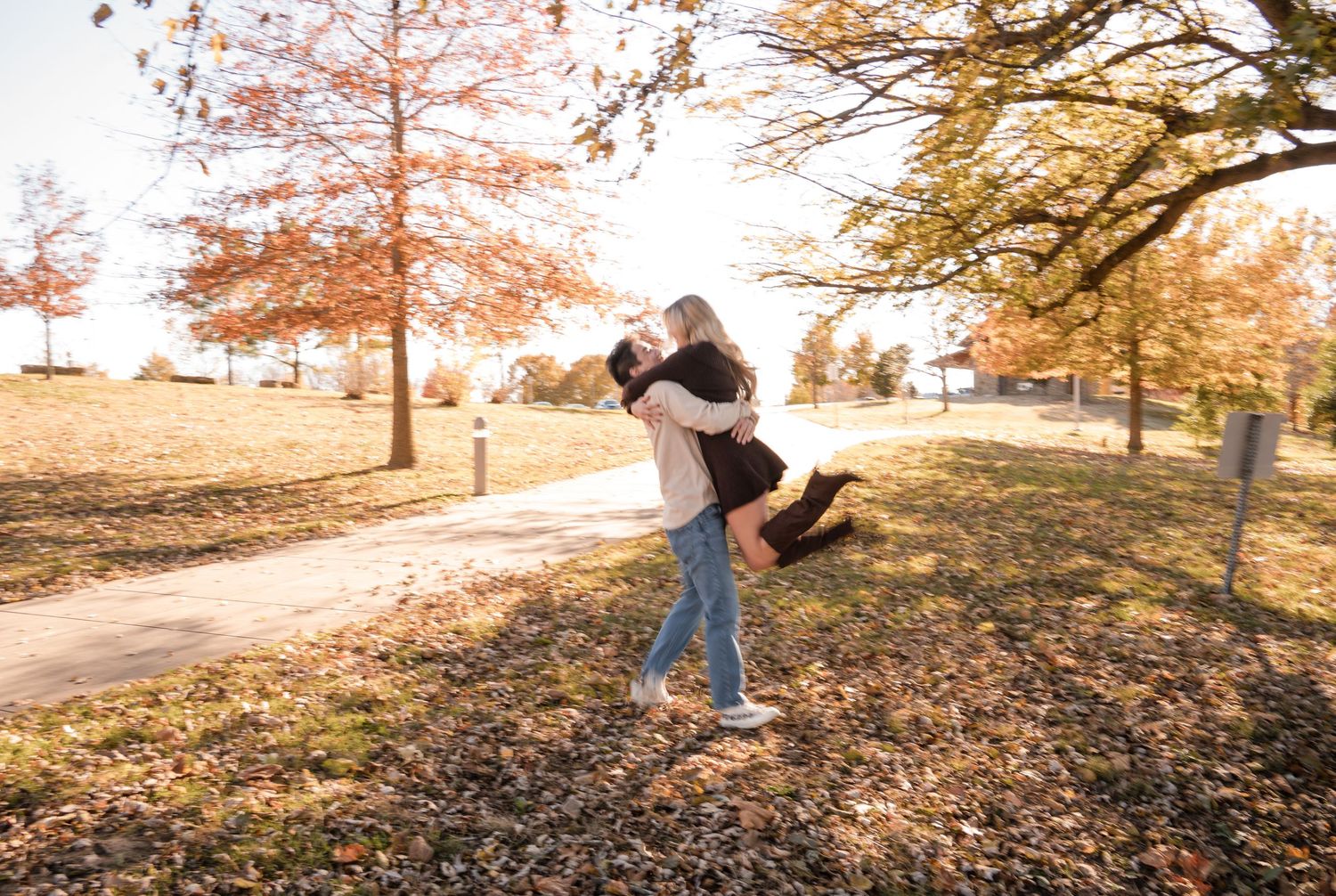 A couple shares a romantic moment in a park during autumn with fallen leaves and golden sunlight.