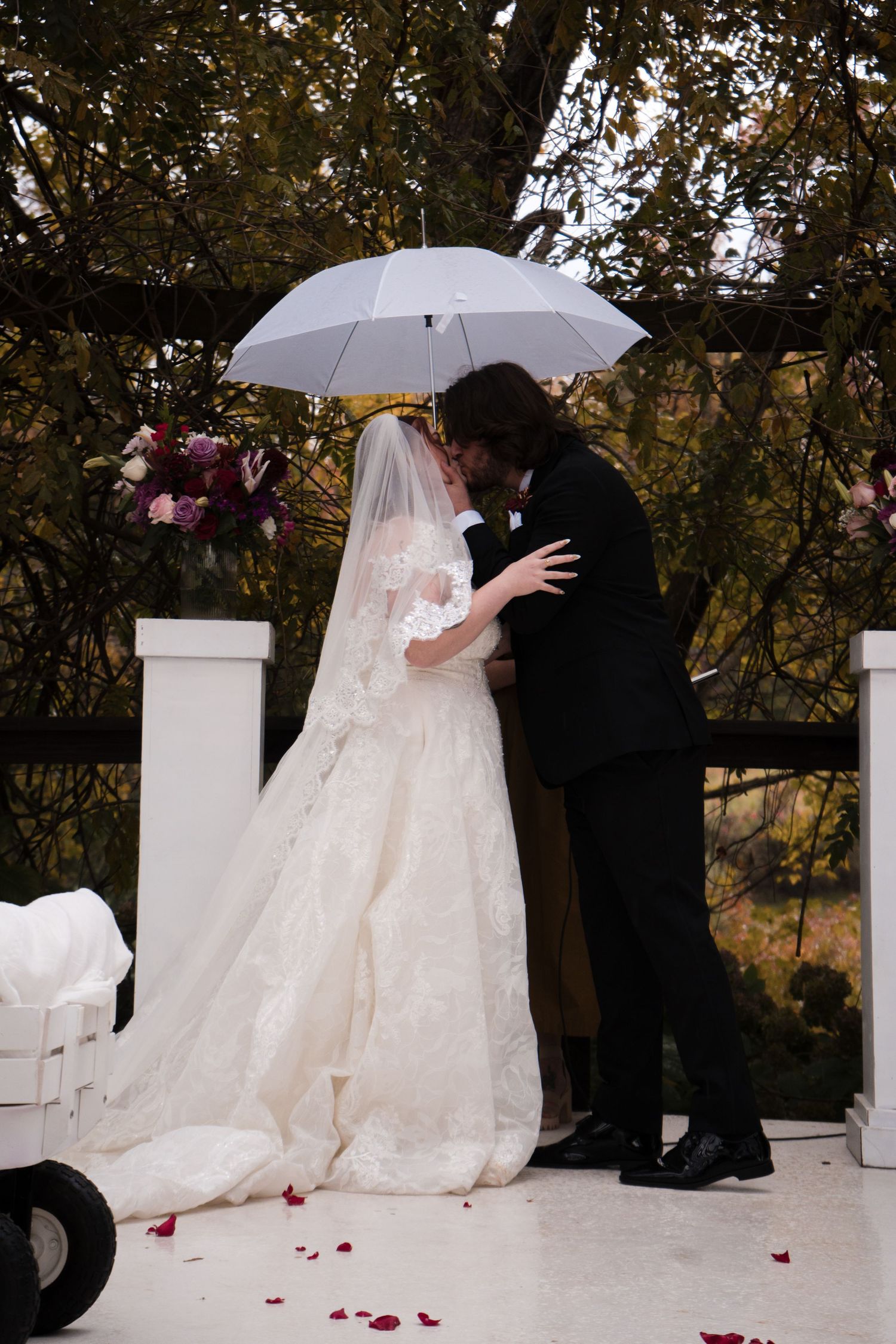 Wedding ceremony under white umbrellas with flowing dress and rose petals scattered on ground.