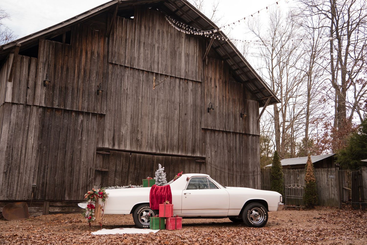 A classic car sits parked next to a rustic wooden barn in a rural setting during autumn.