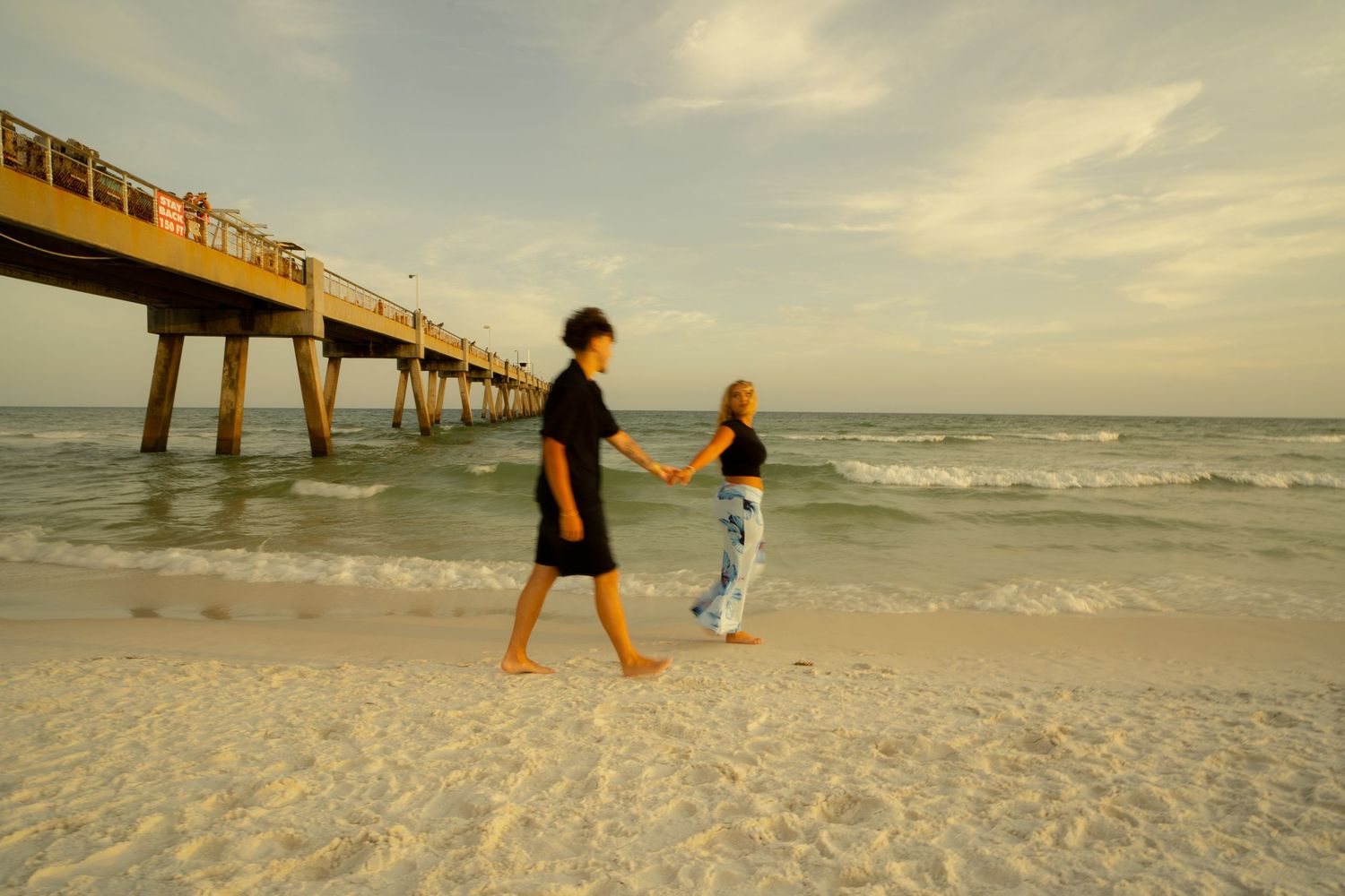 Couple holding hands walking barefoot on sandy beach near ocean pier at sunset.