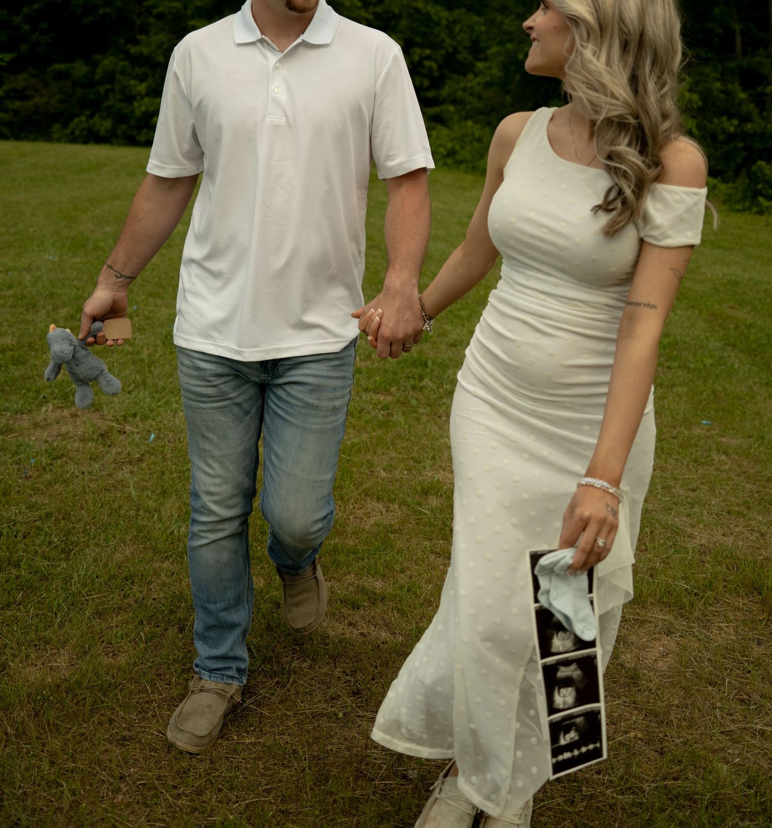 Couple holding hands outdoors, woman in white dress holding ultrasound photos, man holding blue toy, pregnancy announcement.