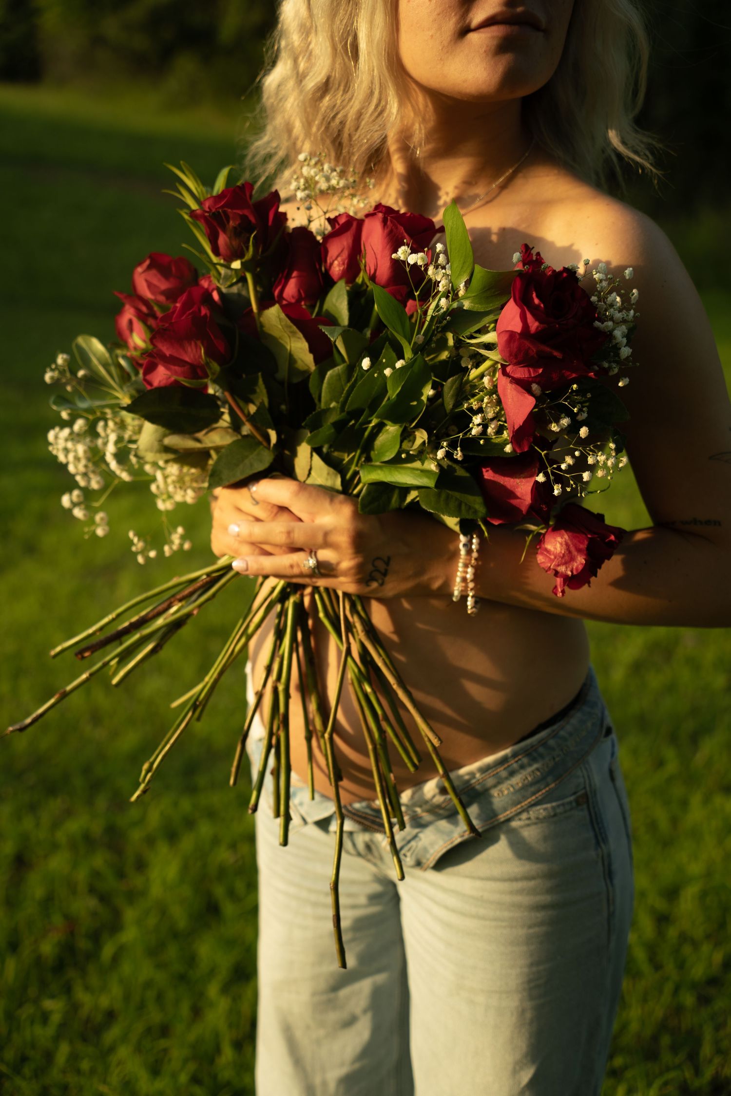 Woman holding bouquet of red roses and baby's breath outdoors in golden hour sunlight, wearing light blue jeans.
