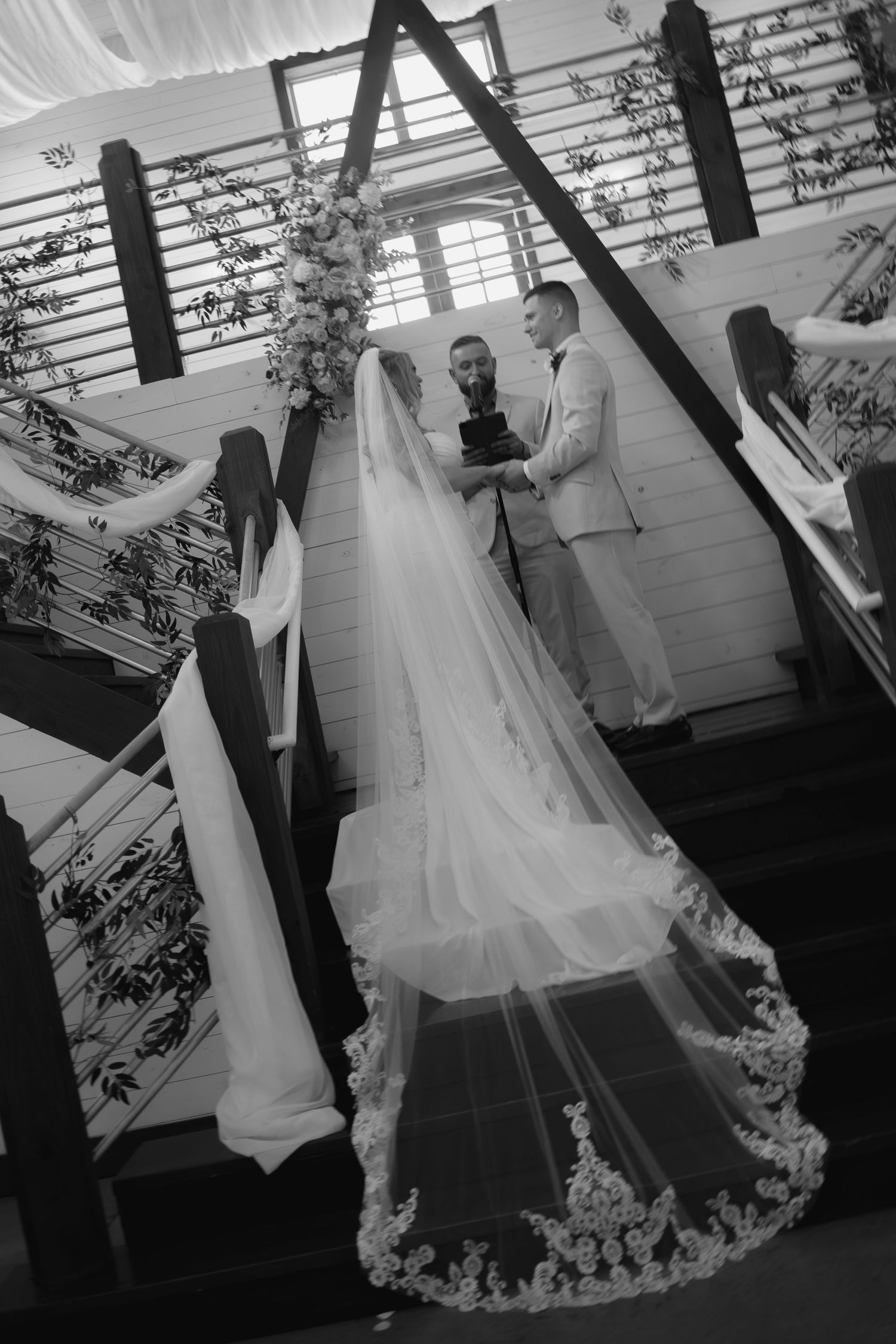 Black and white wedding ceremony photo showing bride in long lace veil and groom exchanging vows on elegant staircase.