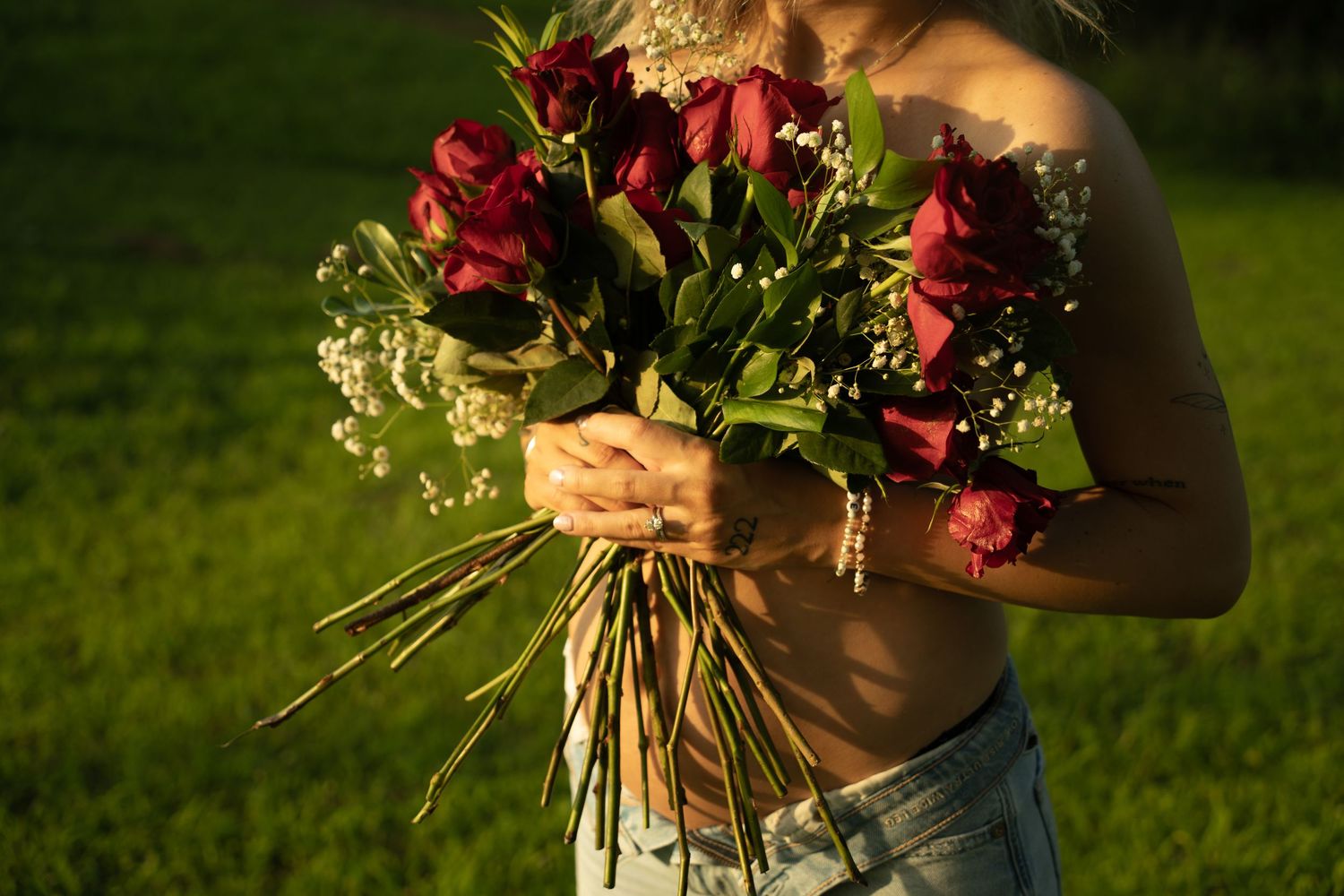 Person holding a bouquet of red roses and white flowers outdoors in a sunny green garden.
