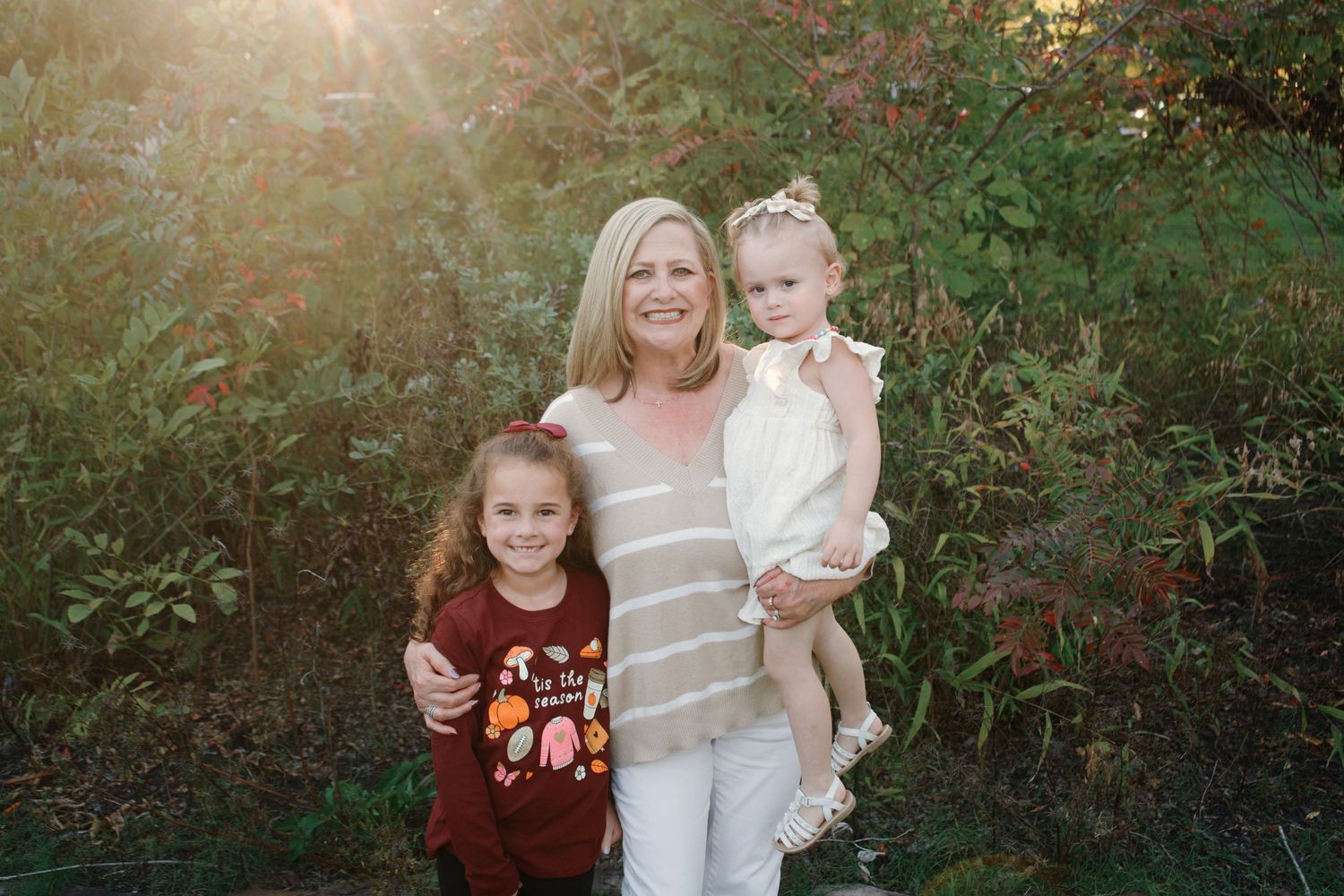 A family poses together outdoors during golden hour in a wooded setting with sunlight filtering through trees.