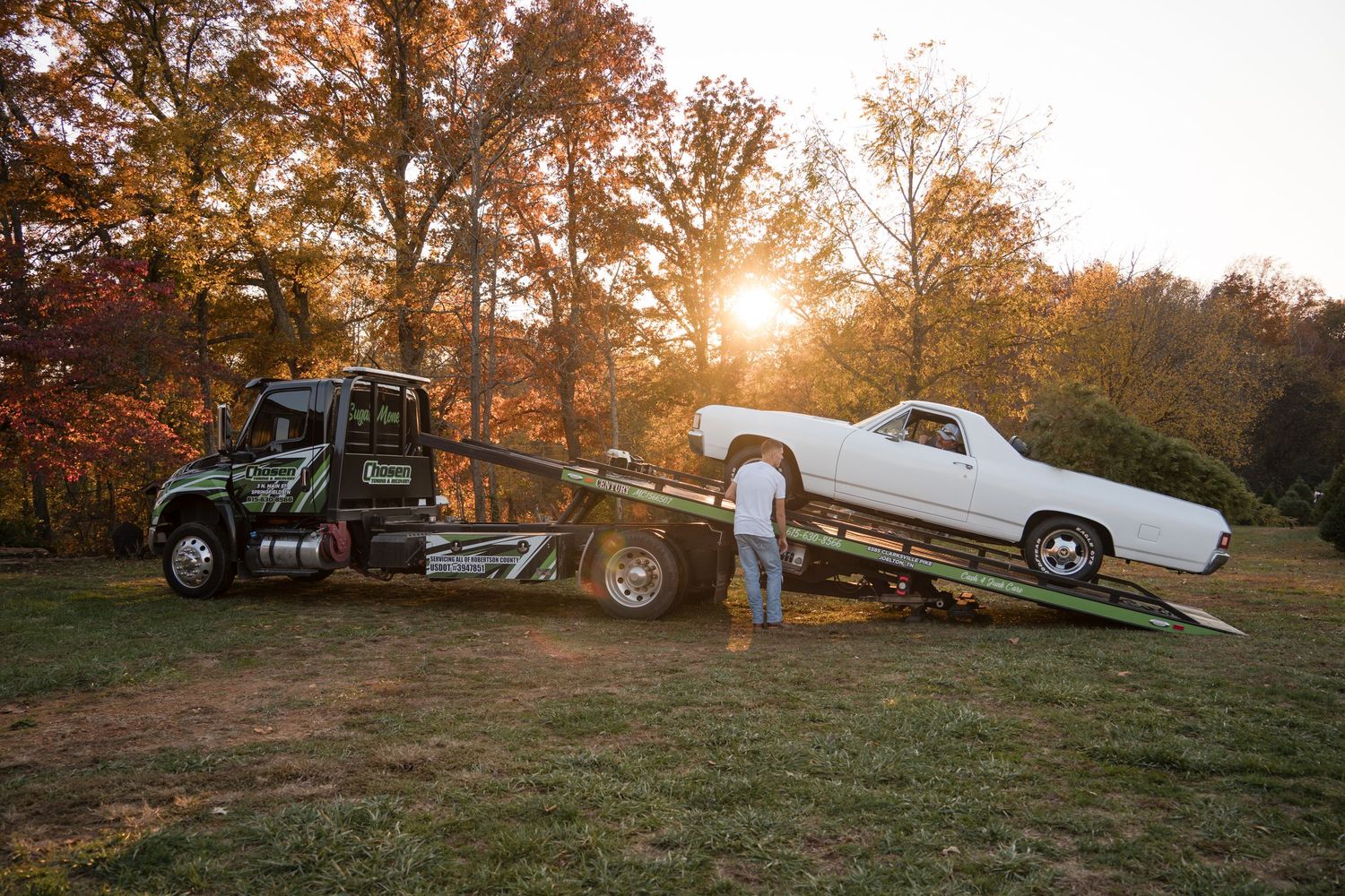 Classic cars and a tow truck are arranged on a grassy hillside during golden hour sunset in autumn.