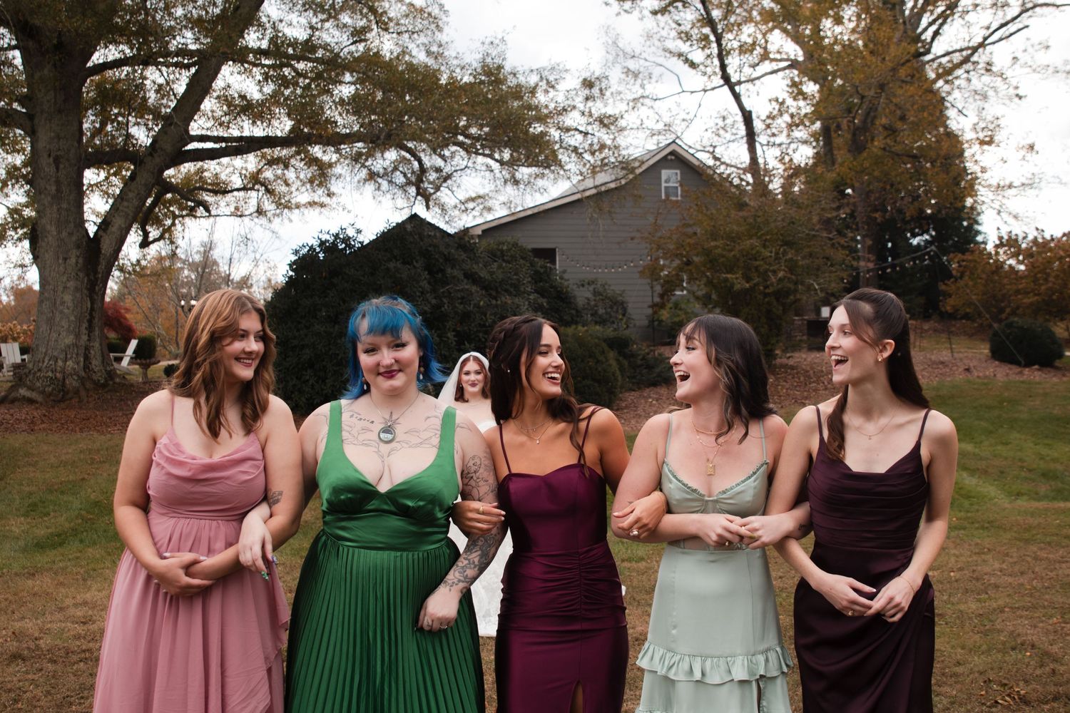 Bridesmaids in pink, green, and burgundy dresses laughing together outdoors.
