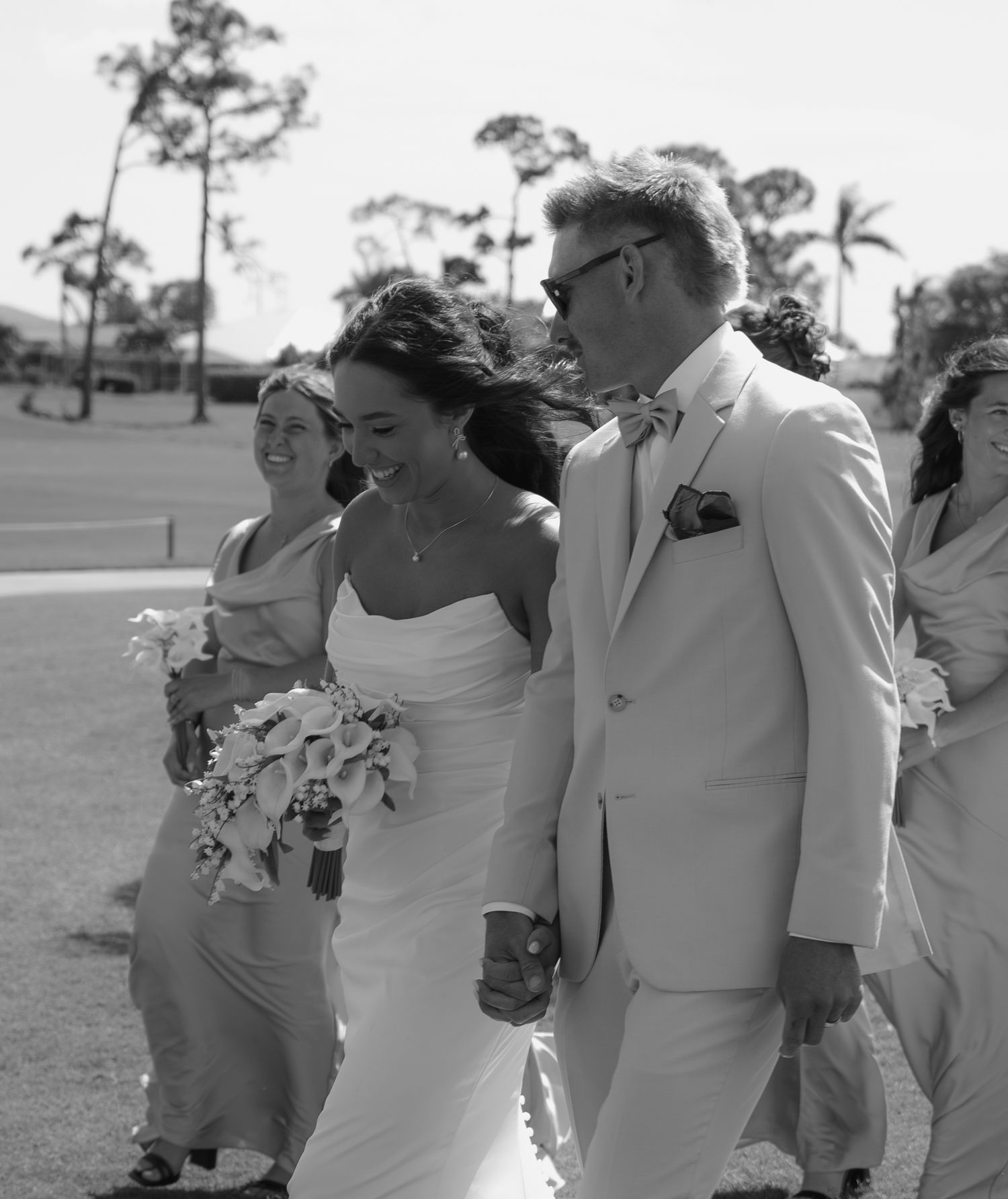 Black and white photo of a smiling bride and groom in a light suit holding hands, walking with bridesmaids outdoors.