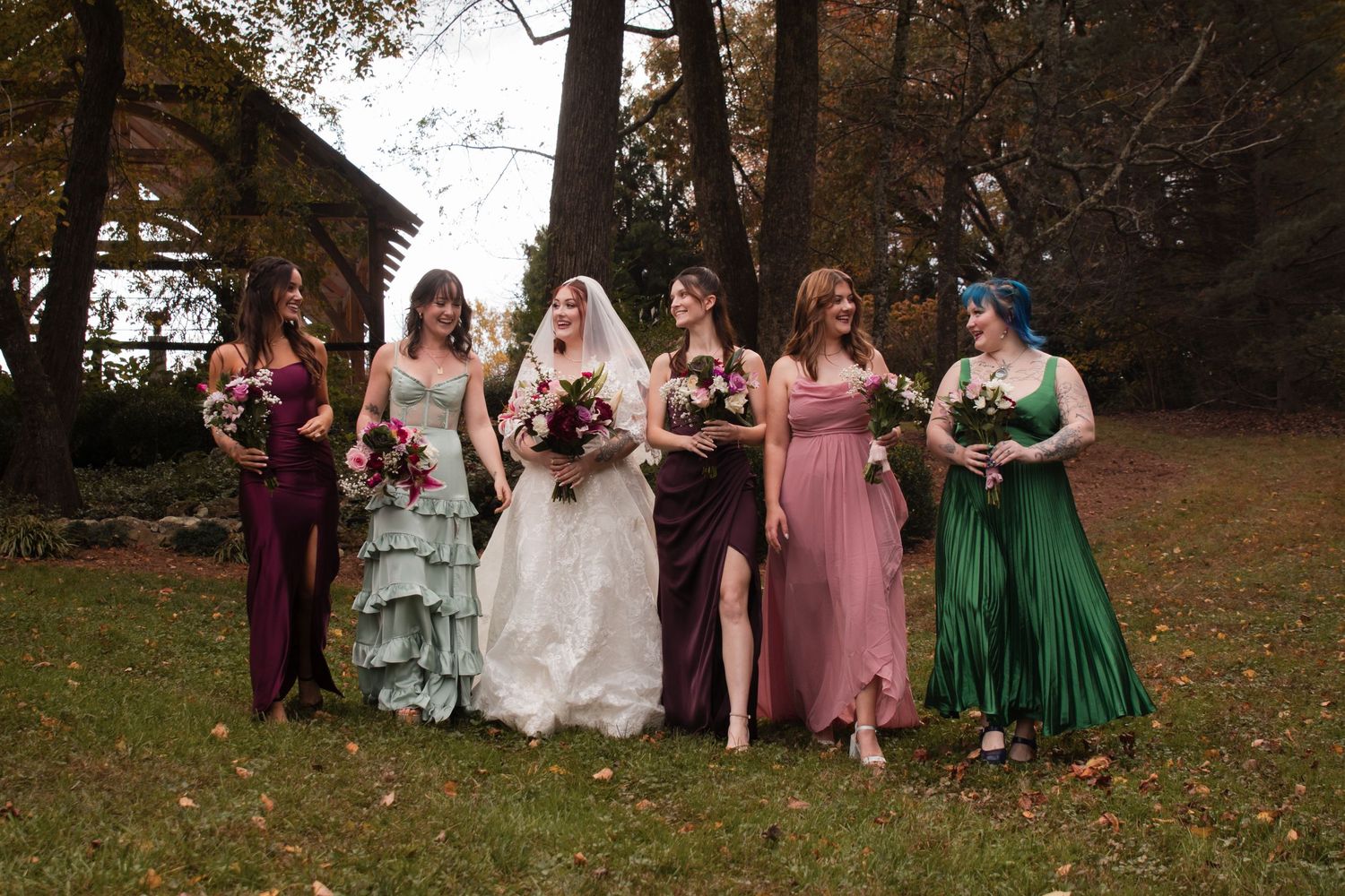 Wedding party in colorful dresses poses together on autumn grounds with fallen leaves.